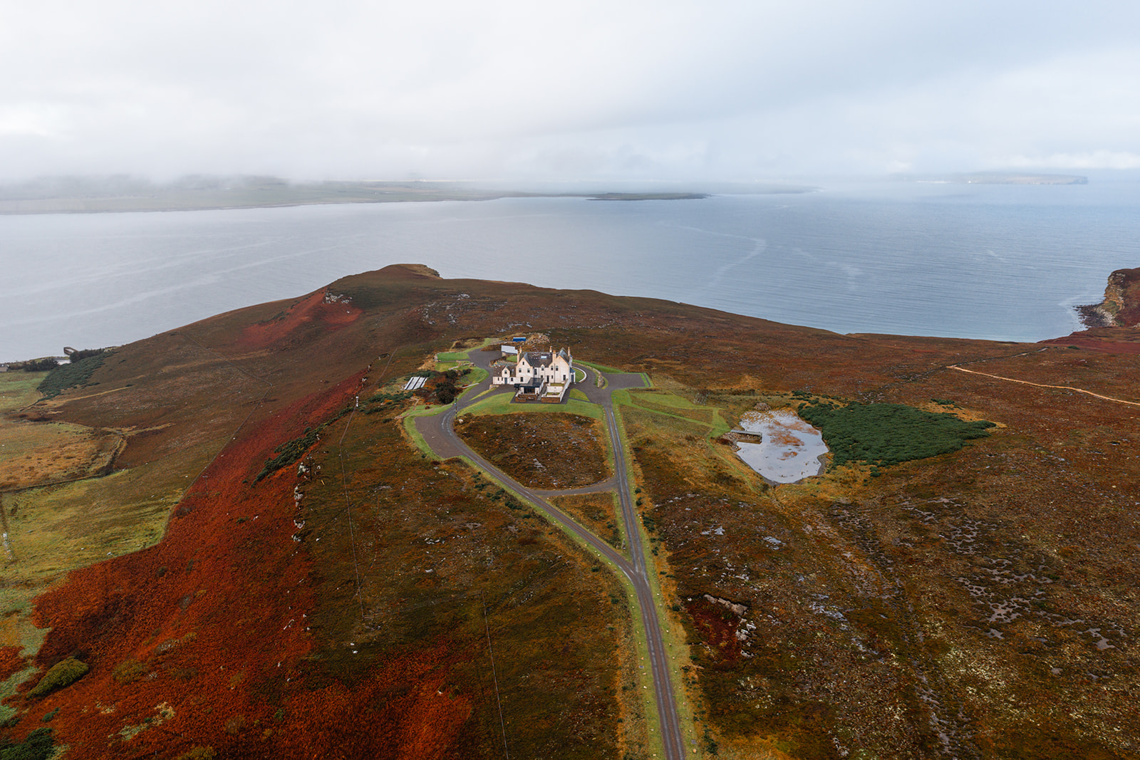 An aerial view shows a white mansion on a headland above the sea, a winding road crossing red autumn moorland.