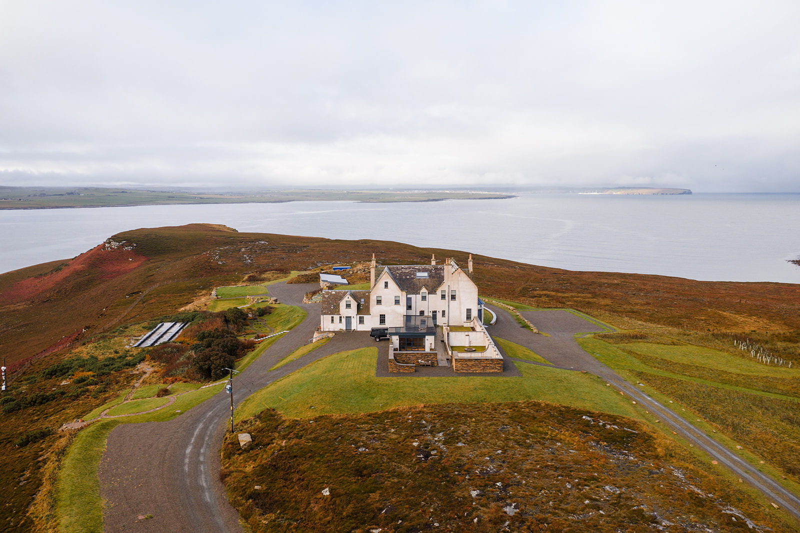 An aerial view shows a white mansion on a grassy headland above a wide sea loch, a road winding through heather-covered moorland.