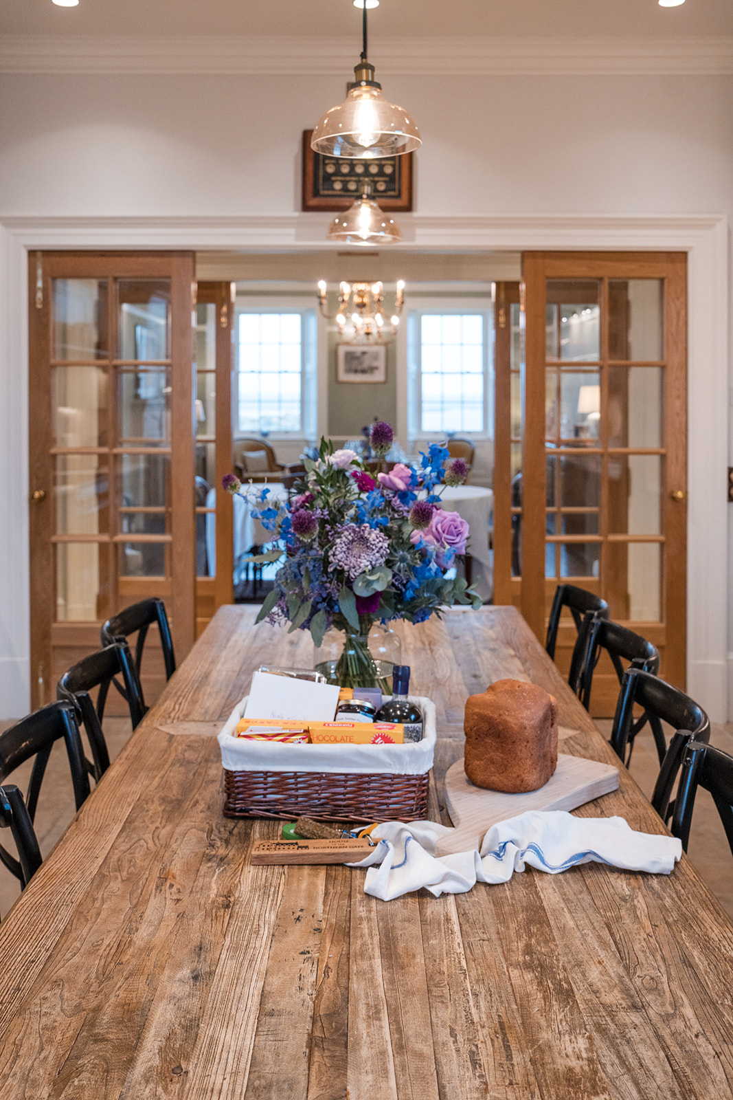 A vase of purple and blue flowers stands on a long wooden table set with a bread loaf and hamper, glazed doors opening onto a dining room beyond.