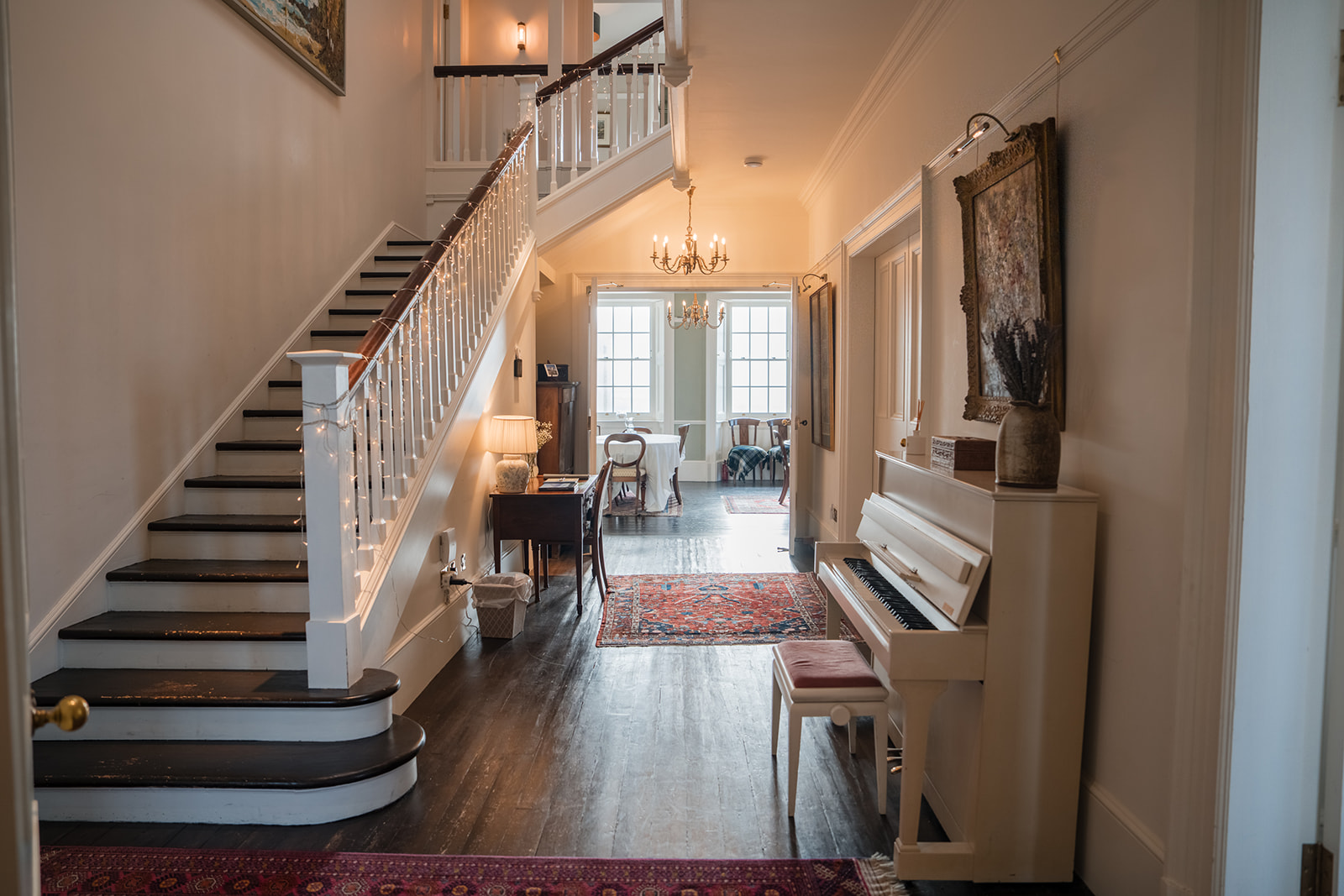 A grand staircase with a white balustrade rises beside a white upright piano, a chandelier and armchairs visible in the room beyond.