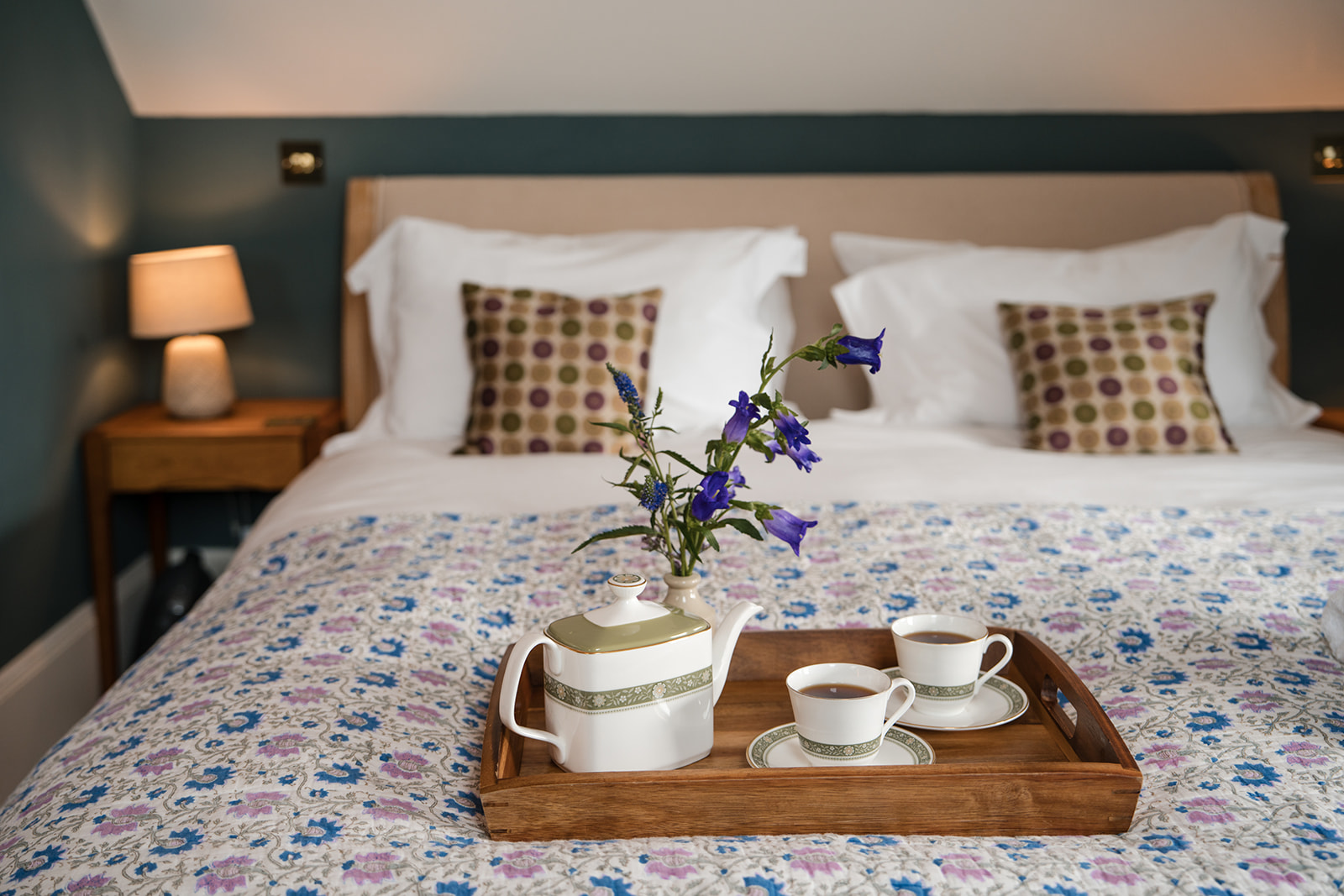 A wooden tray holding a teapot and two cups sits on a floral bedspread, a vase of bluebells and pillows with patterned cushions behind.