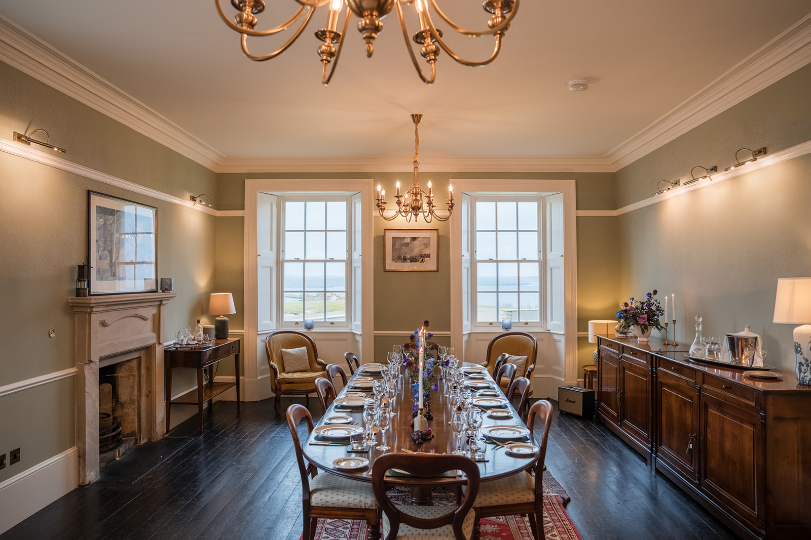 A long dining table set for a formal meal stands beneath a brass chandelier, a fireplace and sideboard along the walls.