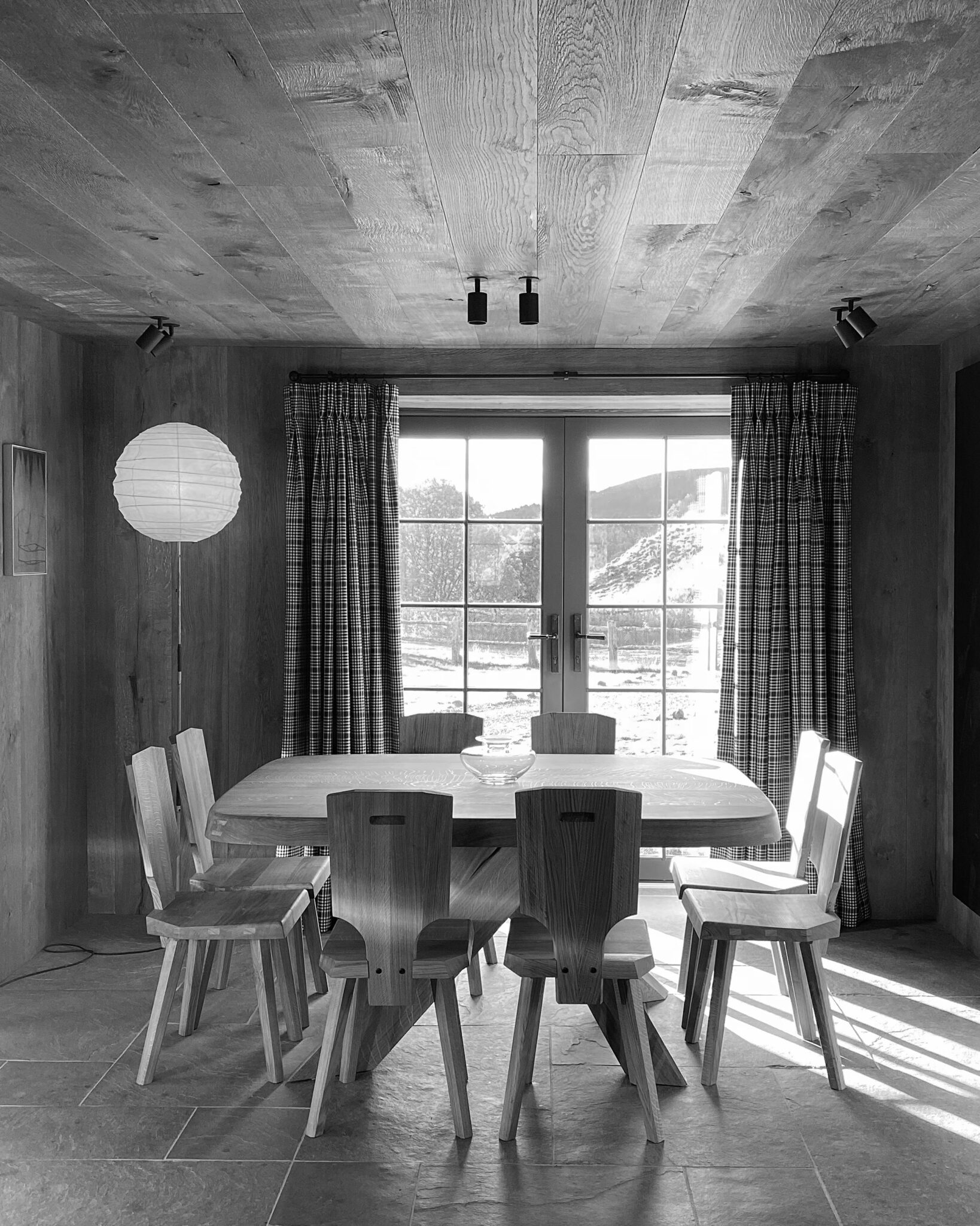 A wooden dining table with chunky chairs stands before glazed doors dressed in tartan curtains, a paper lantern light and framed print to the side.