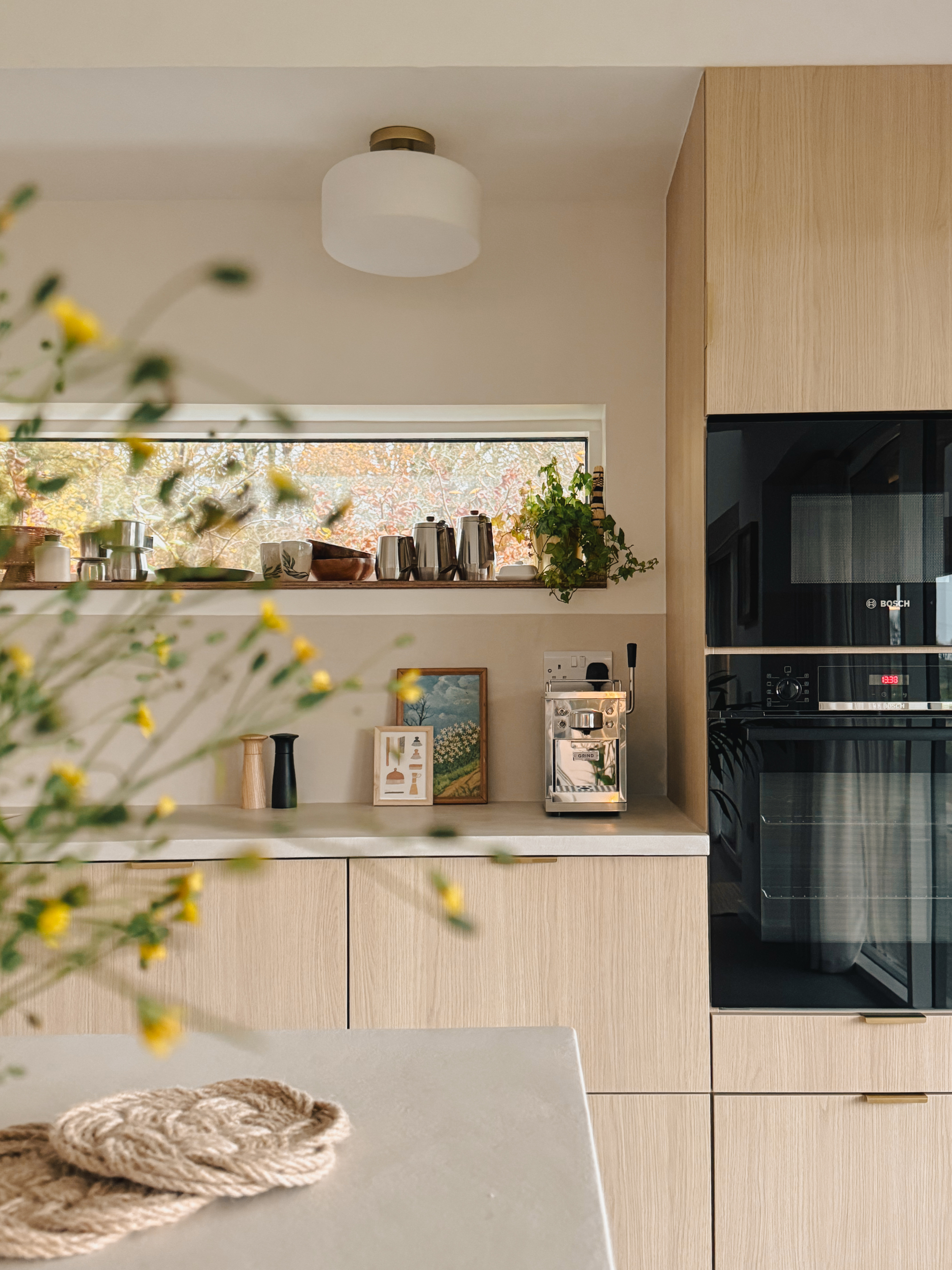 A built-in oven stands beside open shelving displaying a small painting and vintage coffee pots, wildflowers blurred in the foreground.
