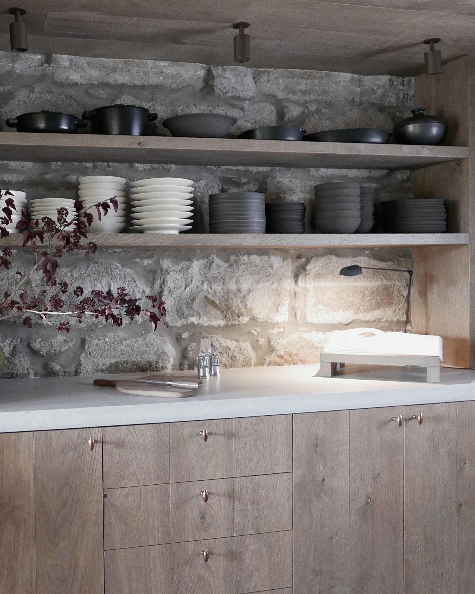 Open shelving holds dark cookware and stacked plates above a stone wall, a marble worktop with a chopping board and salt cellar below.