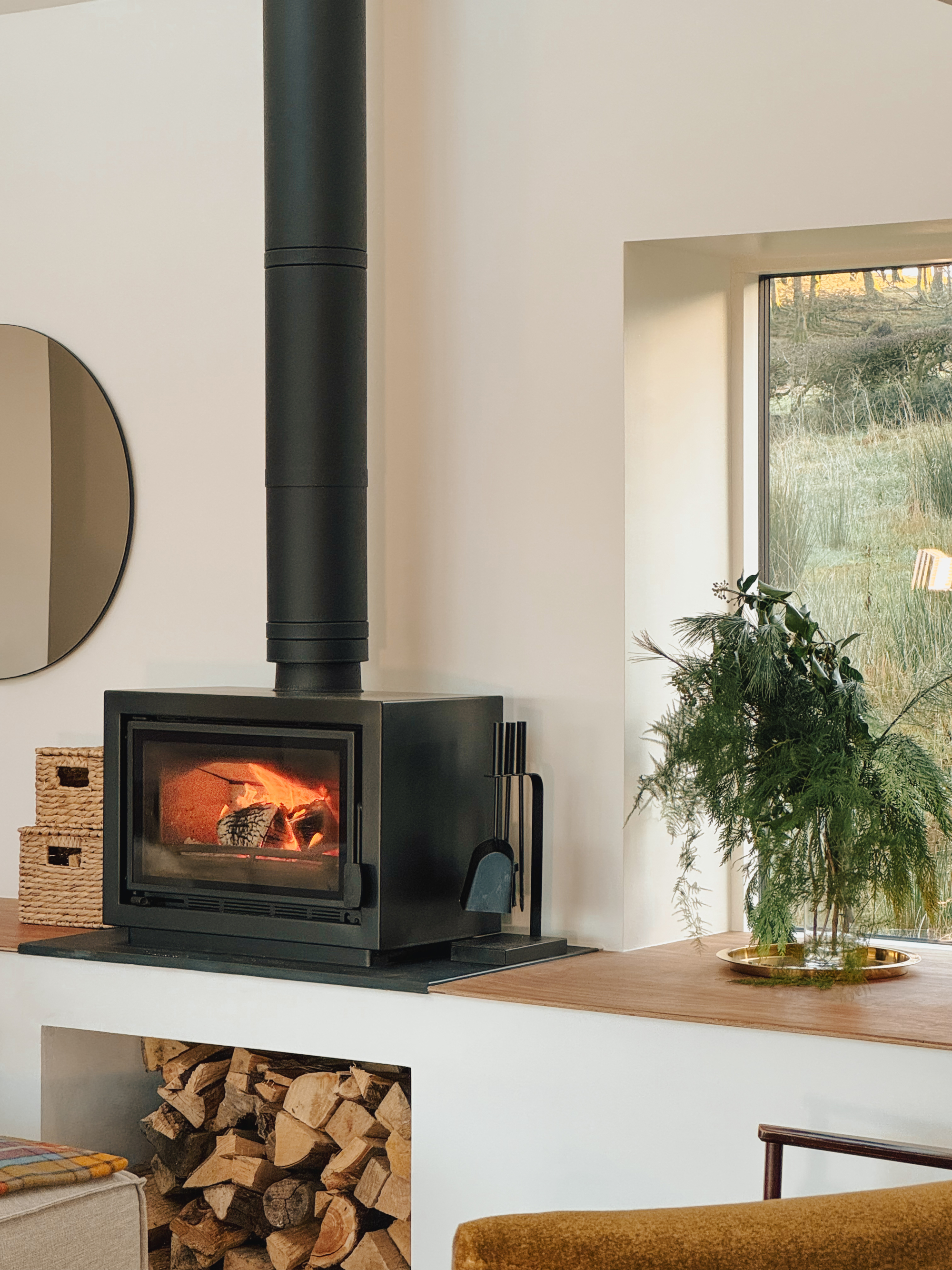 A wood burning stove burns brightly beneath a tall flue between a round mirror and a window onto grassland, a stack of woven baskets and logs nearby.