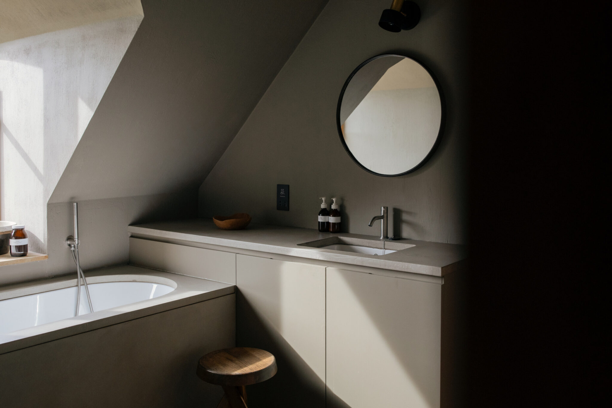 A round mirror hangs above a bathroom counter with a basin, a bath with a wooden stool to the left beneath a sloped ceiling.