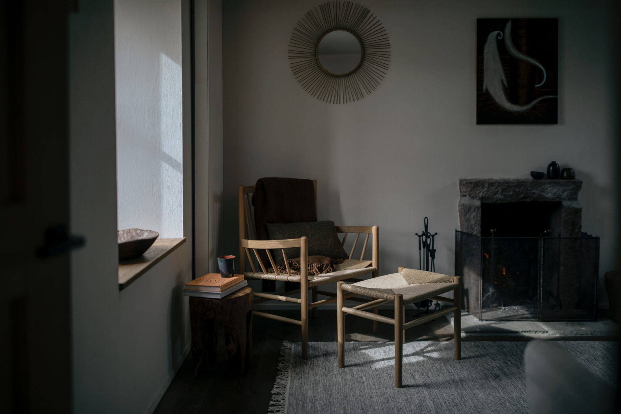 A woven-seat armchair and matching footstool stand before an old stone fireplace fitted with a mesh guard, a round inlaid table in the foreground.