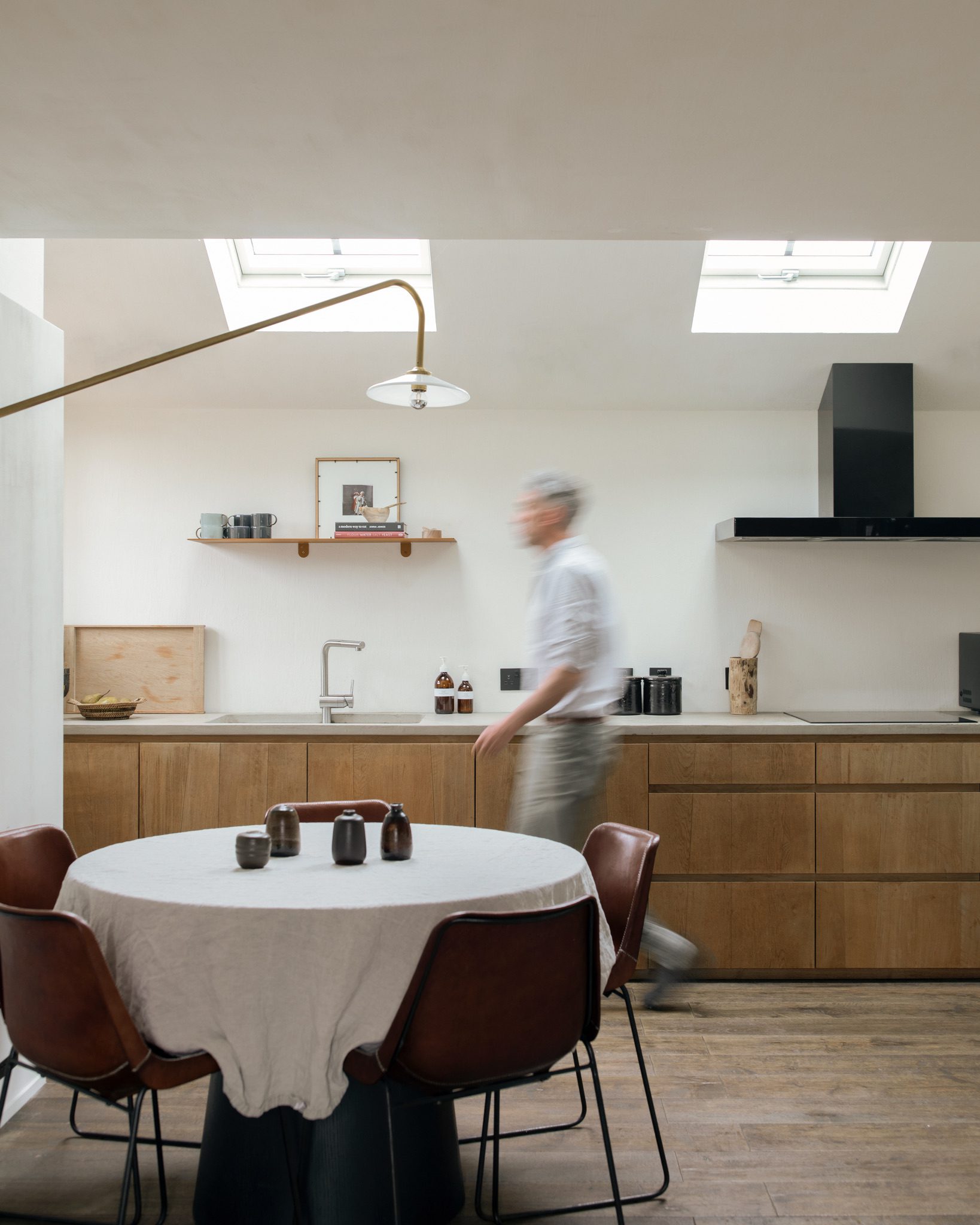 A man walks through a kitchen with wooden cabinetry, a round table set with mugs in the foreground beneath skylights.
