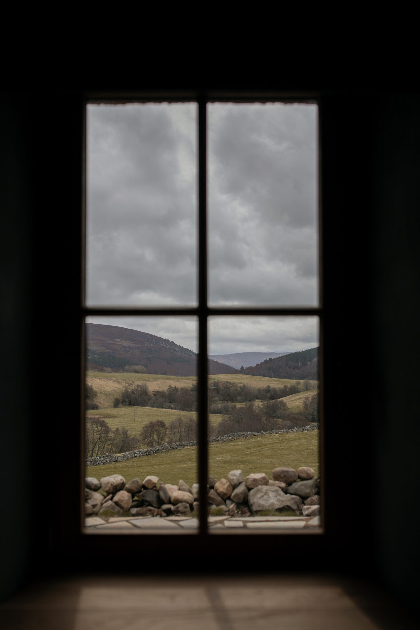 A four-paned window looks out over rolling hills and a dry stone wall.