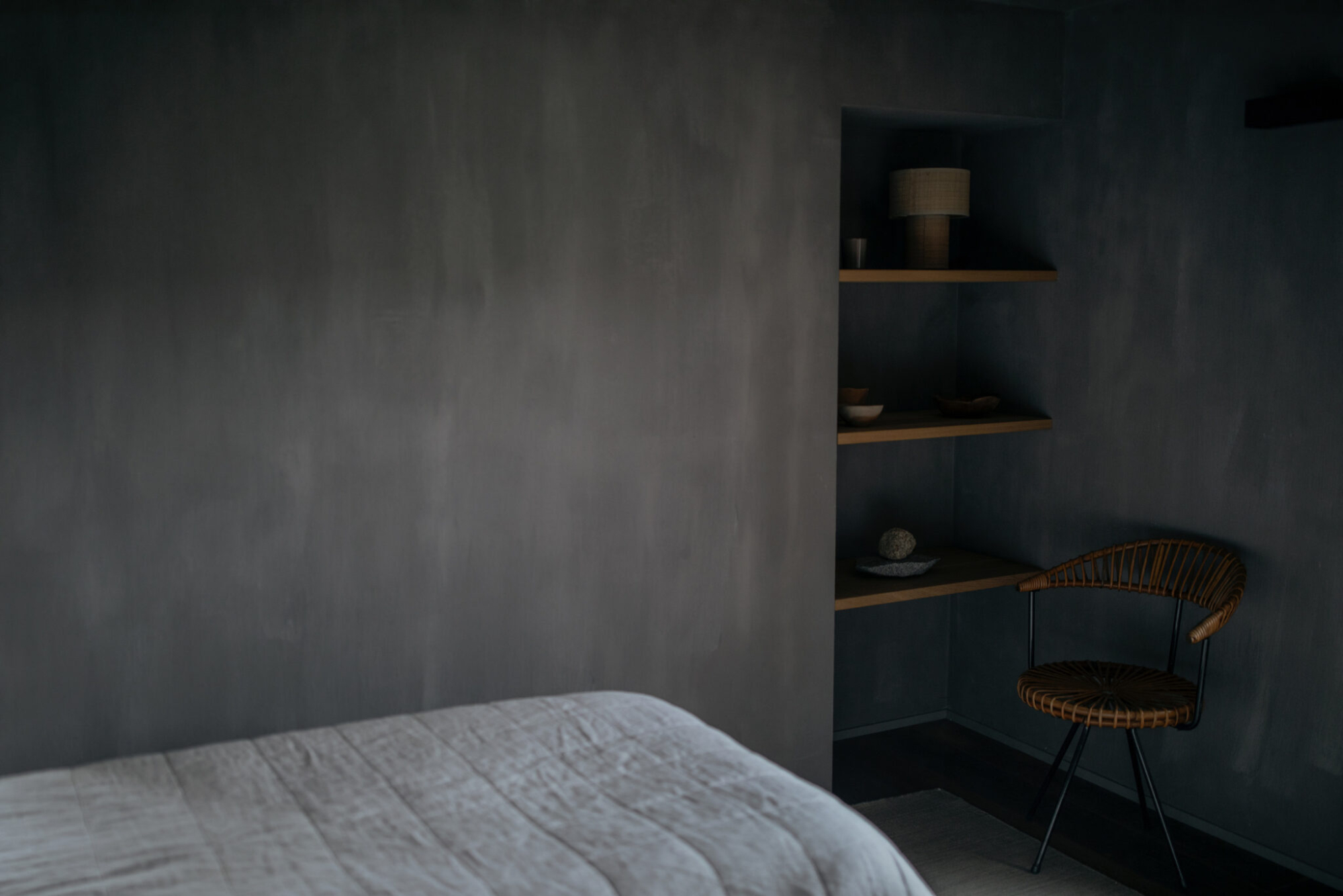 A built-in shelf holds a lamp, small bowls and ornaments in a grey plastered alcove beside a woven chair.
