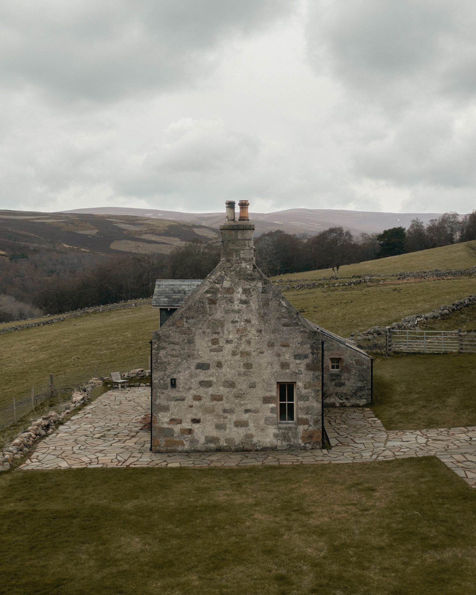 A stone cottage with twin chimneys sits on a grassy hillside, dry stone walls crossing the fields around it.