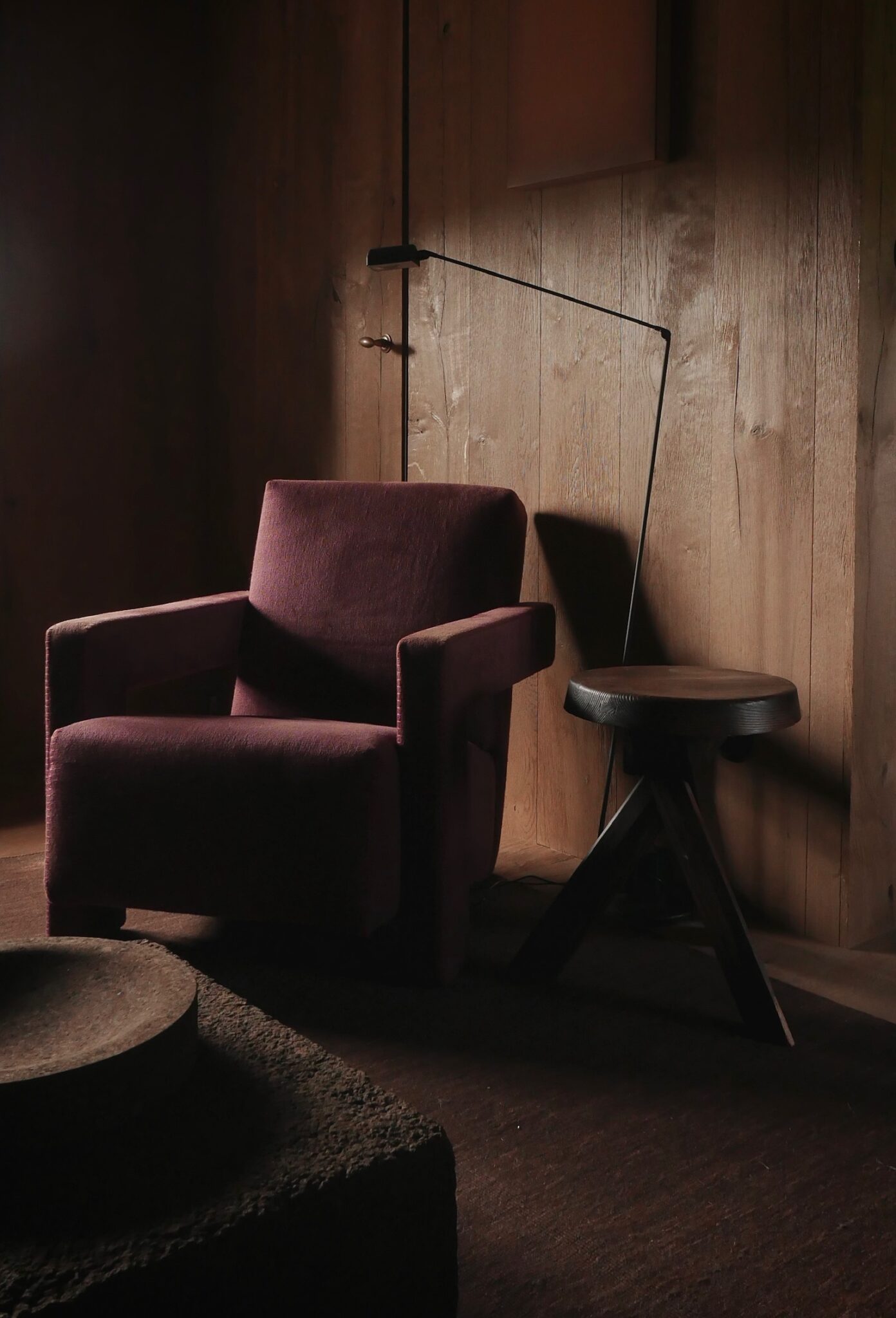 A burgundy armchair sits beside a round wooden stool and an angled floor lamp against timber panelling.