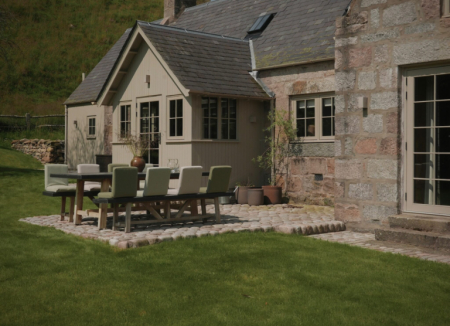 A wooden table and chairs with green cushions stand on a stone patio beside a gabled stone house, a lawn stretching in front.