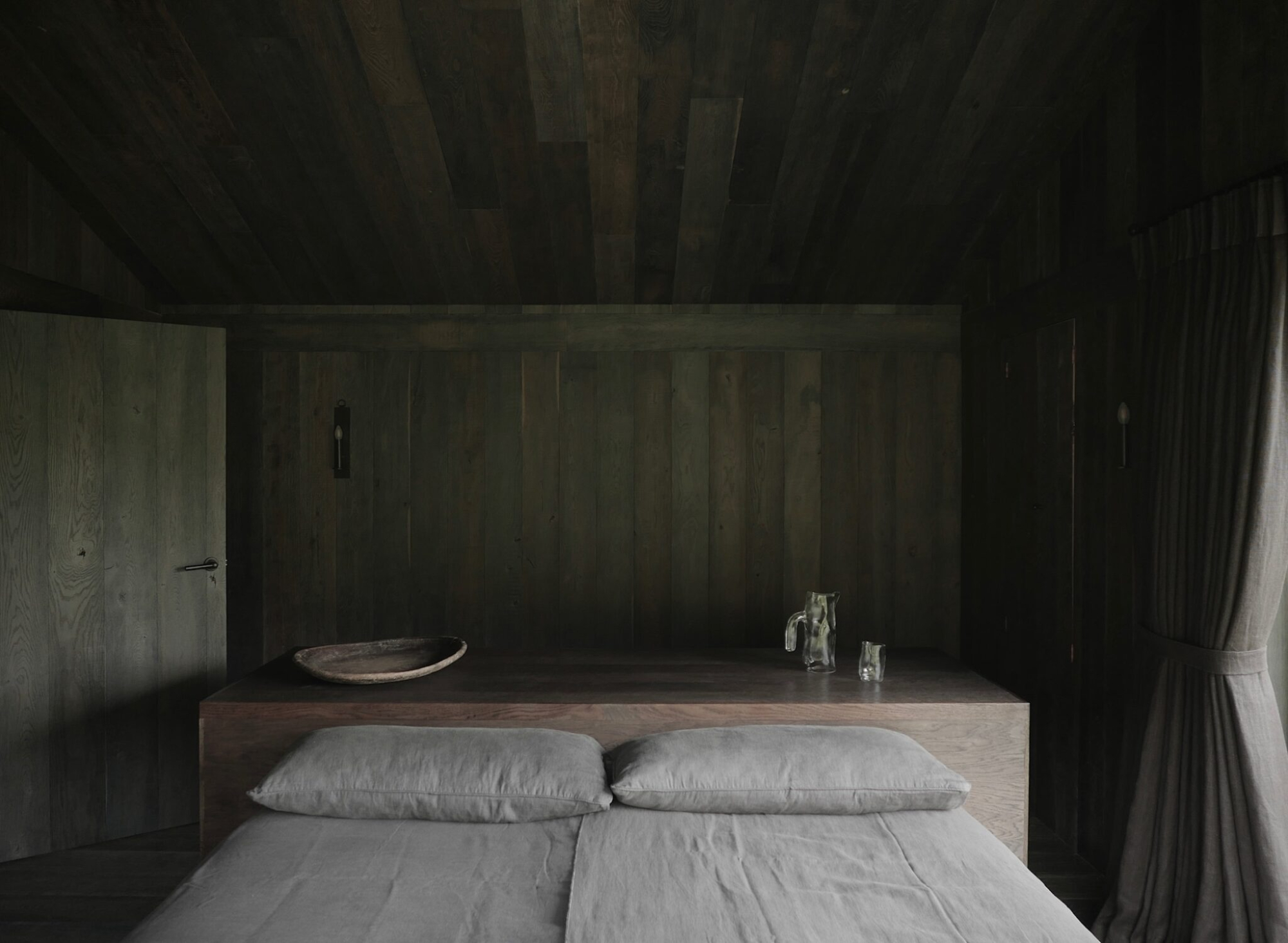 A bed with grey linen sits against dark timber panelling, a round tray with a jug and glasses on the headboard shelf.