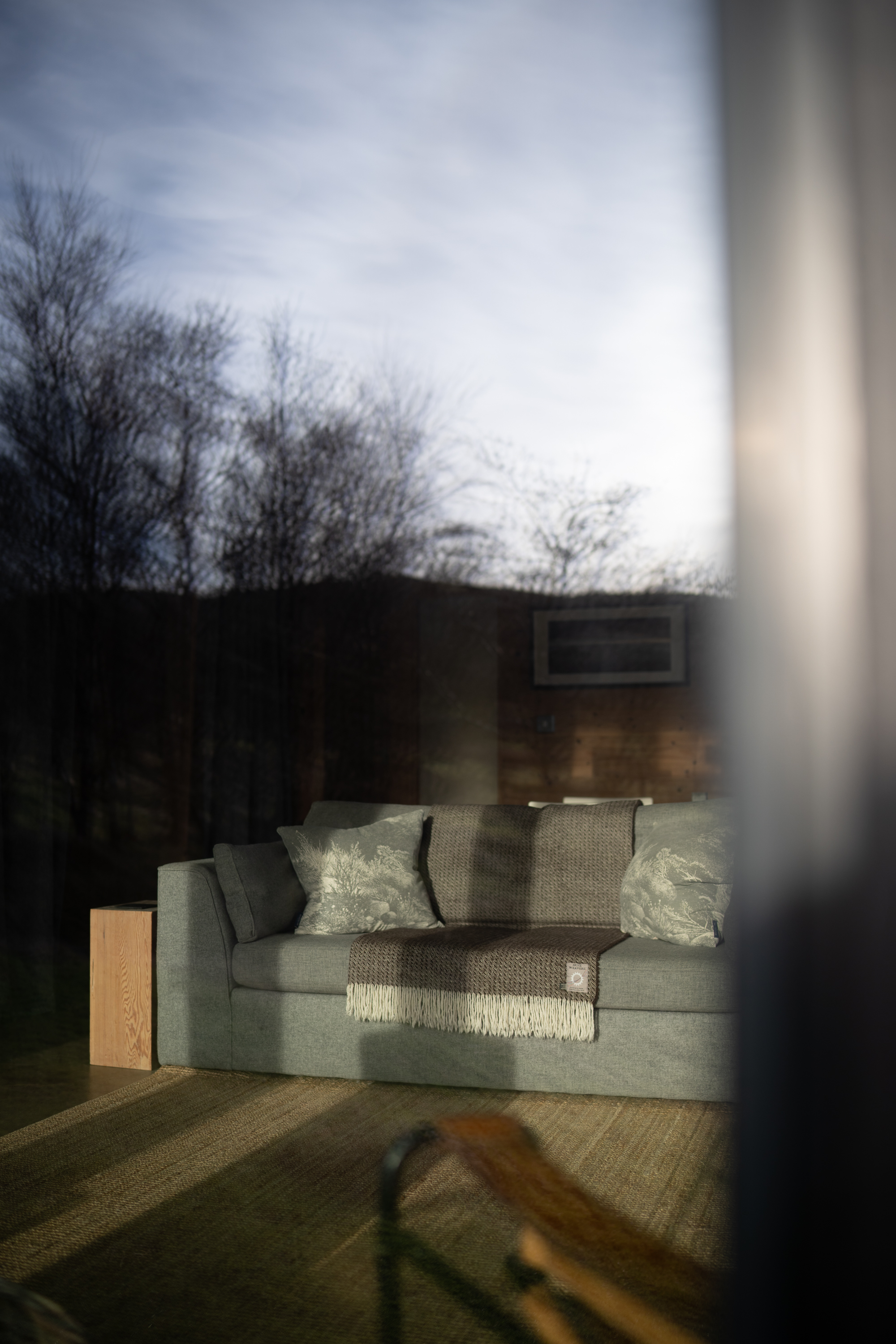 A grey sofa with cushions and a fringed throw is reflected in a window, bare trees and hills visible outside at dusk.