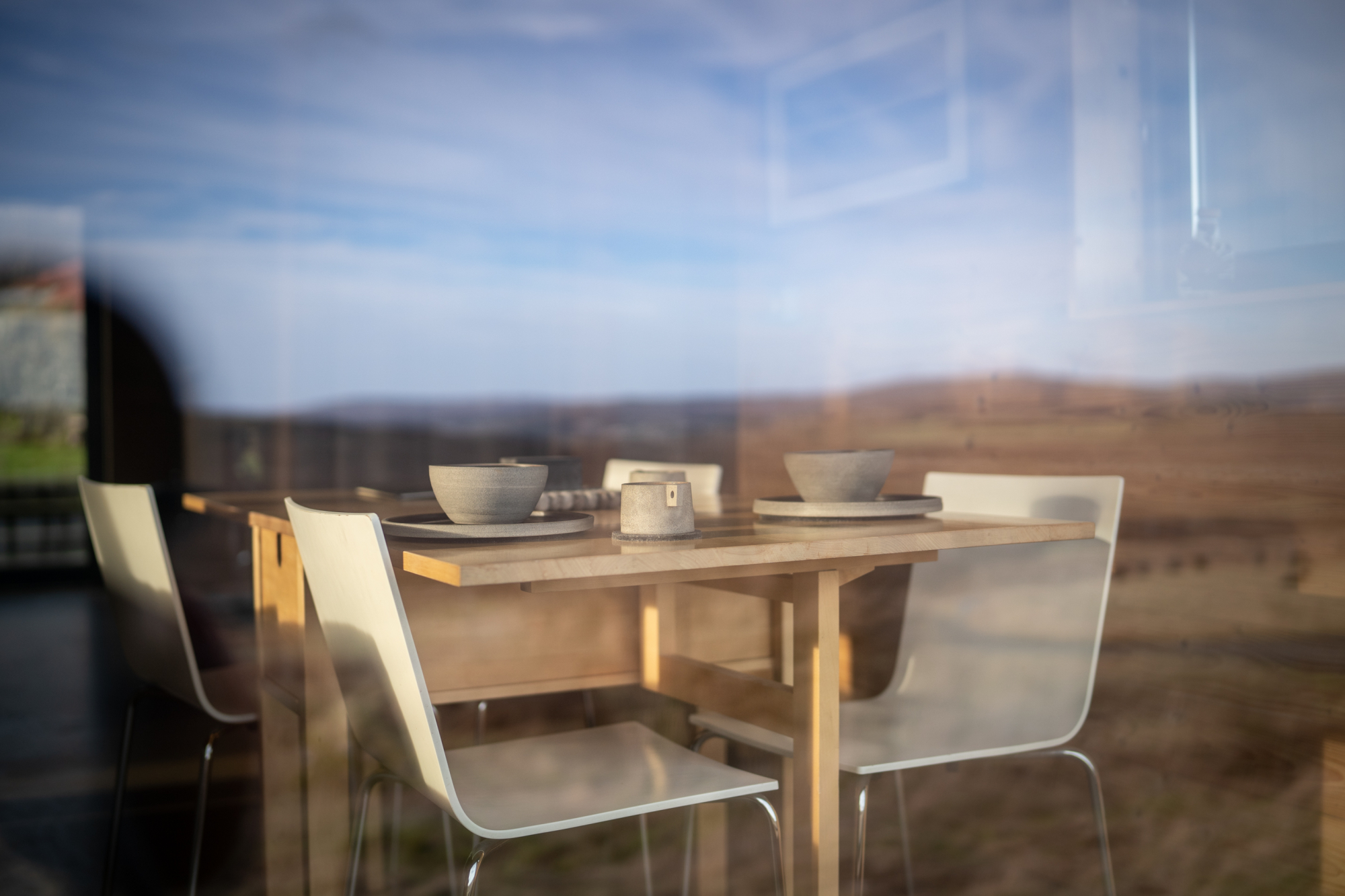 A wooden table set with bowls and a jug stands before a window framing moorland and hills, white chairs around it.