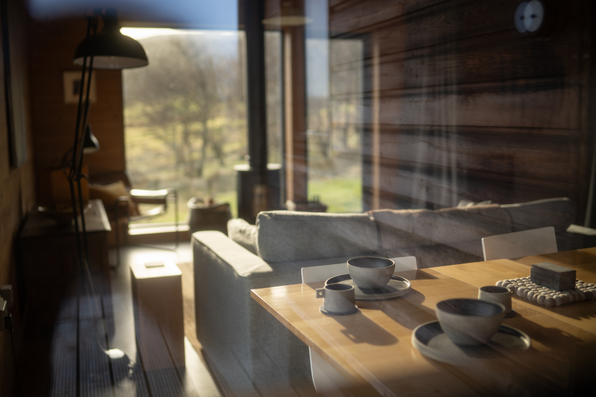 A wooden dining table set with cups and saucers stands before a sofa and wood burning stove, moorland visible through the full-height windows.