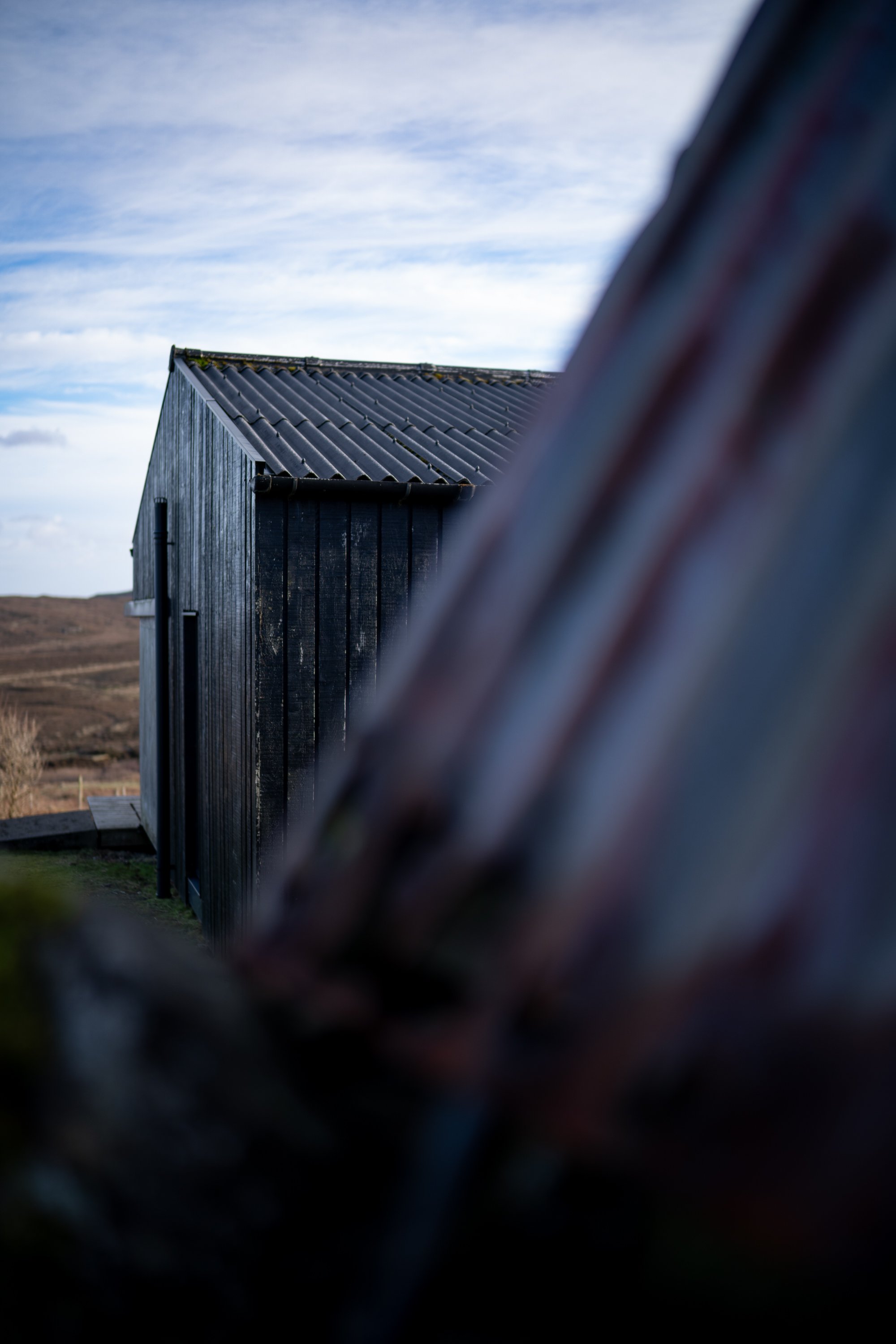 A weathered timber shed with a corrugated roof stands on open moorland, seen past the edge of a rusted corrugated wall.
