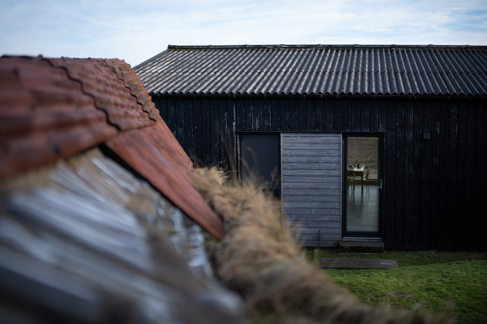 A moss-covered red pantiled roof sits in the foreground of a black timber cabin, its glazed door reflecting a dining table inside.