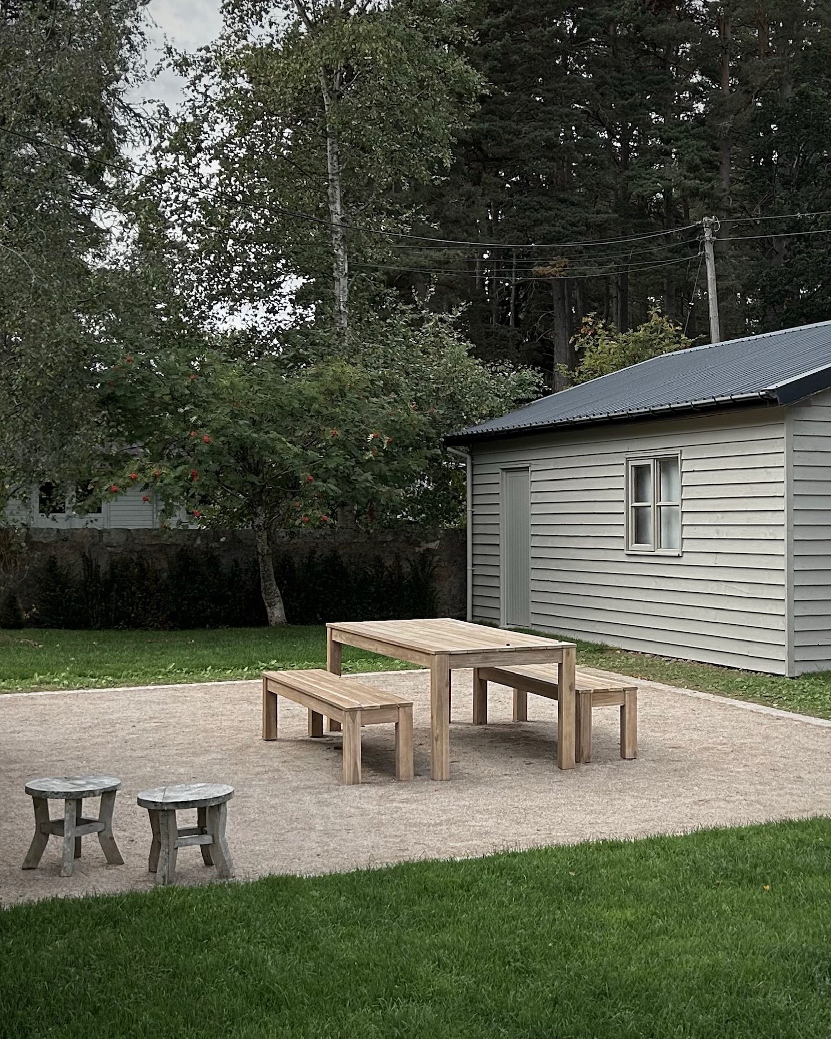 A wooden picnic table and benches stand on a gravel patio before a grey clad outbuilding, trees and a lawn surrounding it.