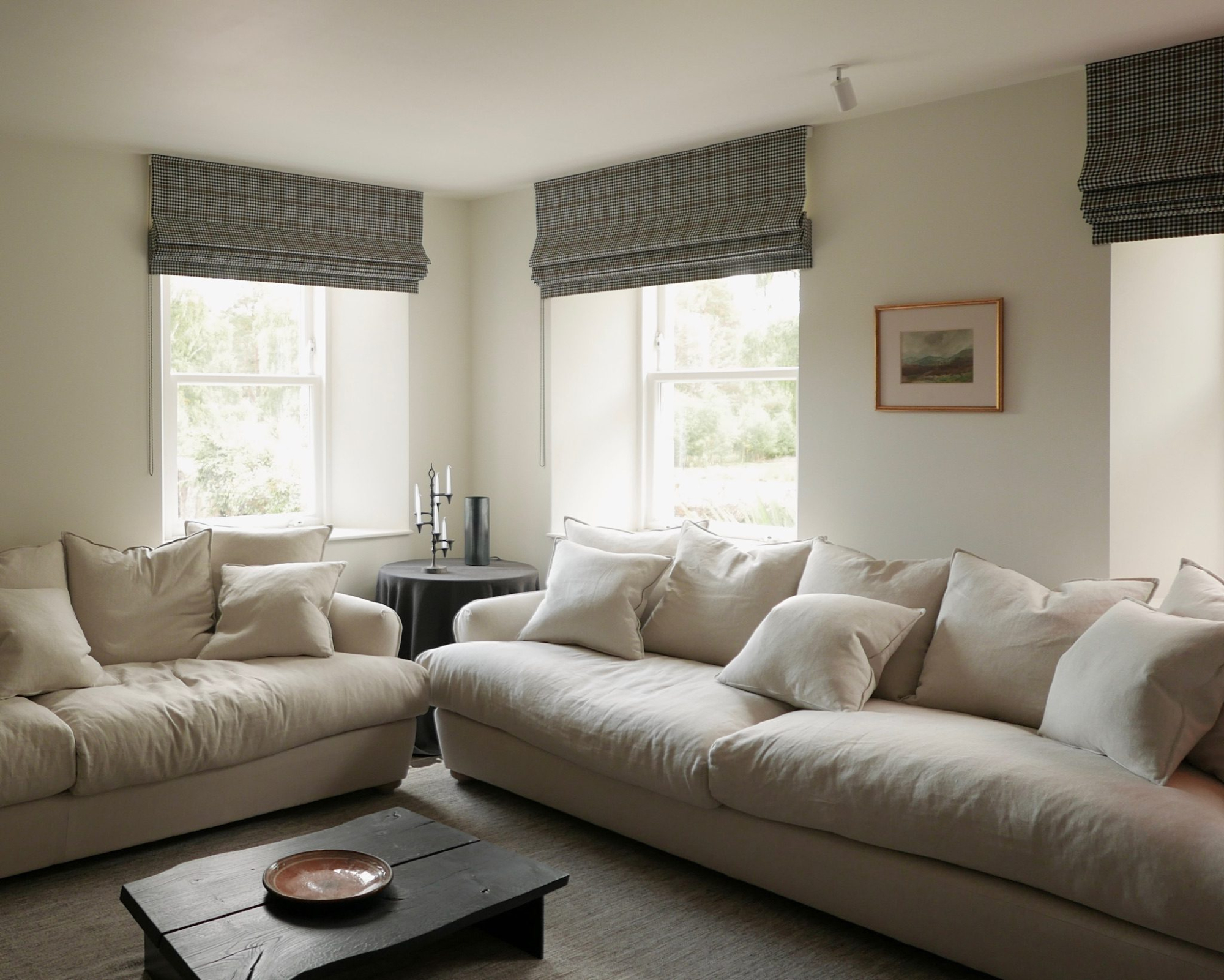 Two grey linen sofas face each other across a dark wooden coffee table, checked roman blinds at the windows and a framed landscape print on the wall.