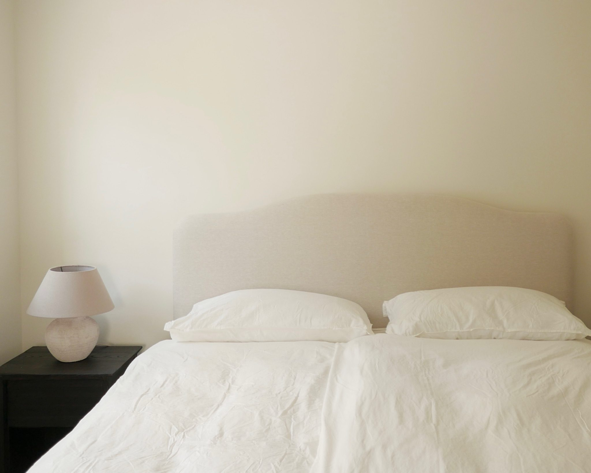 A bed with plain pillows and a taupe upholstered headboard sits beside a round lamp on a dark bedside table.
