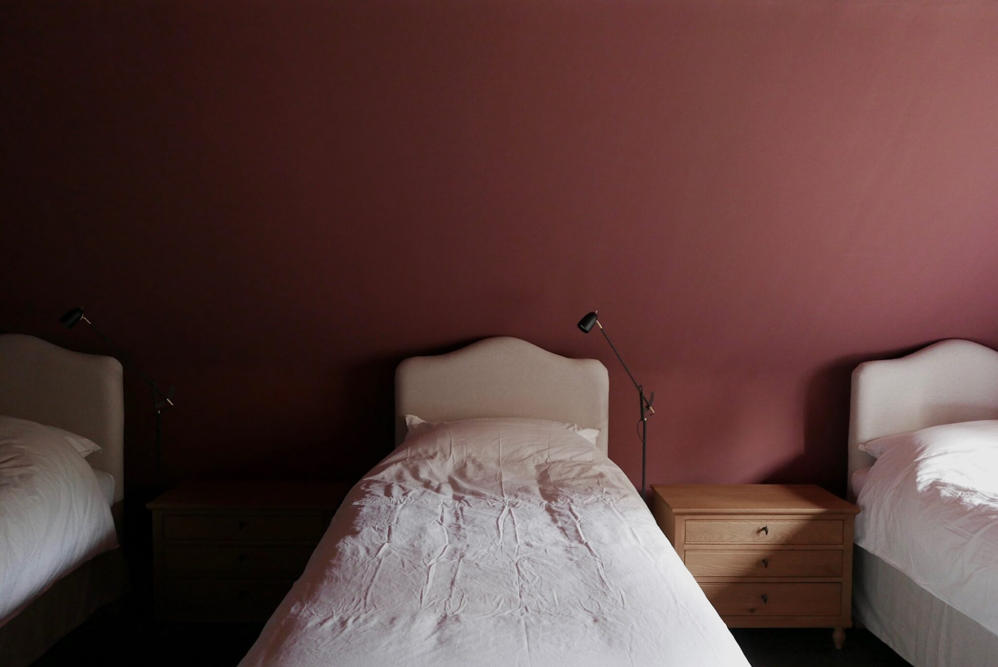 Three beds with taupe headboards stand against a dusky pink wall, a wooden bedside cabinet and wall-mounted reading lamp between them.