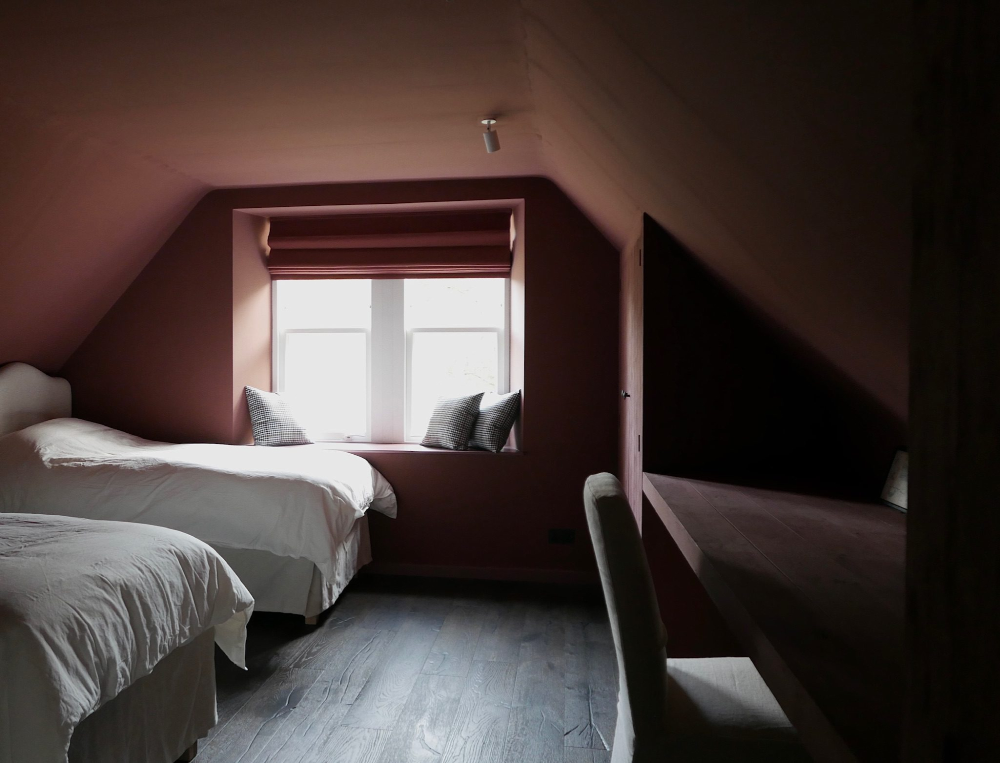 Two single beds with white linen sit beneath a dormer window in a dusky pink room, a desk and chair to the side.
