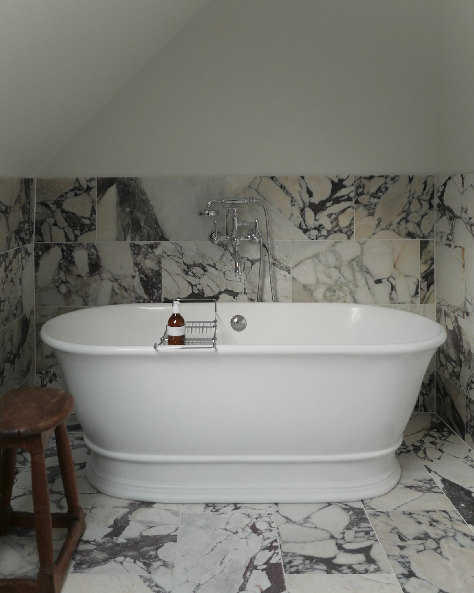 A freestanding bath stands against marble-effect tiling, a wooden stool and a small amber bottle on its rim.