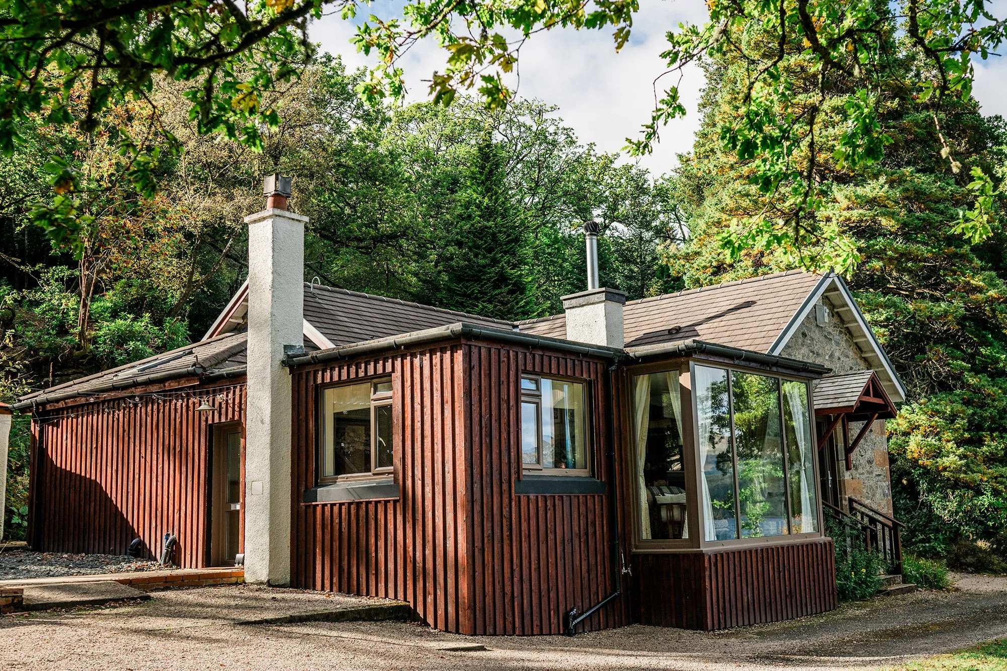 A dark timber-clad extension with a conservatory joins a stone cottage, gravel paths leading around the building through trees.