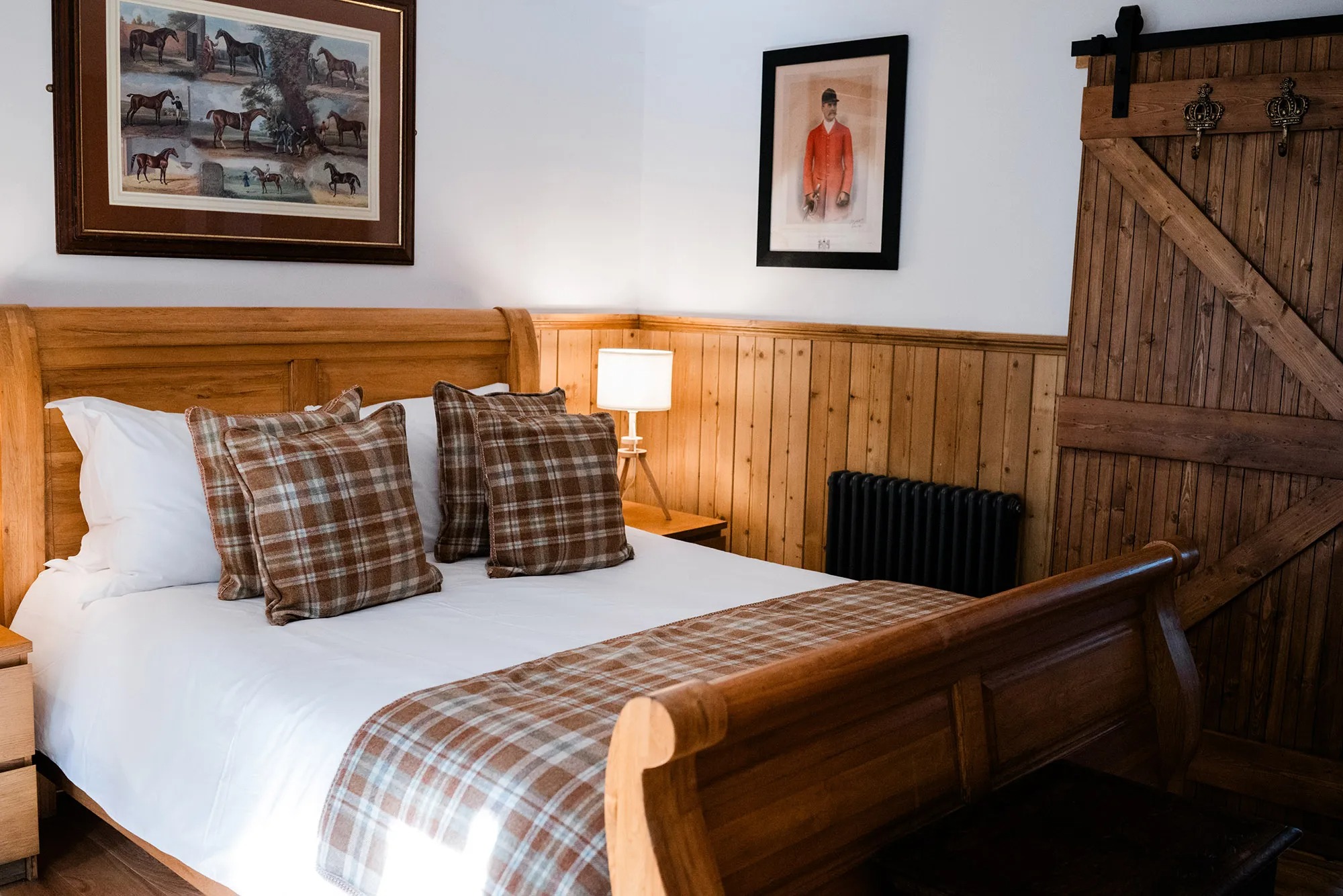 A wooden sleigh bed with tartan cushions sits before panelled walls hung with equestrian prints, a barn door and radiator to the right.