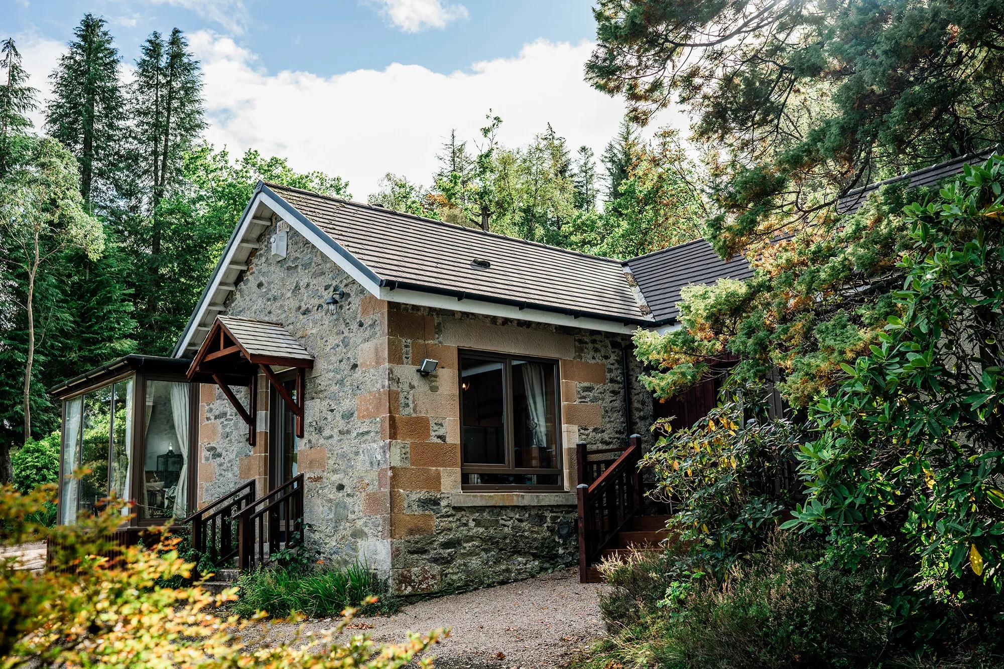 A stone cottage with a gabled porch and conservatory sits among trees, autumn foliage framing the entrance.