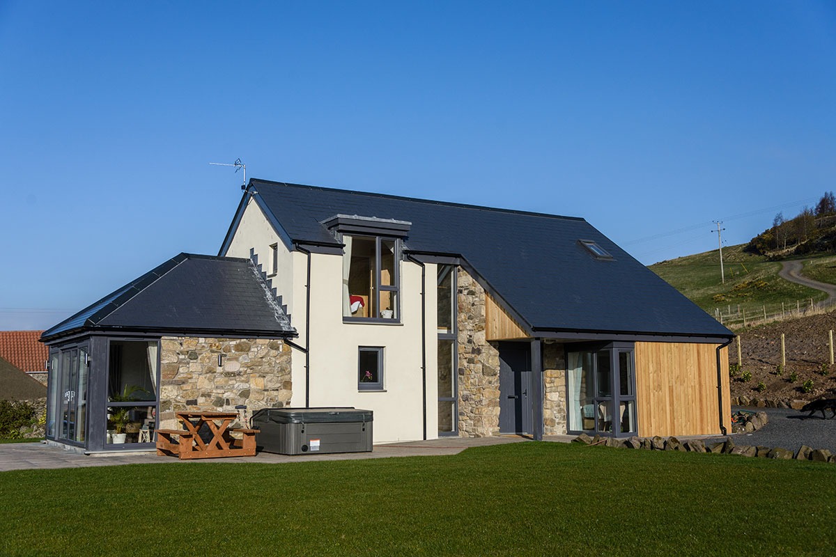 A stone and timber house with a dark slated roof and roof windows sits on a lawn, a hot tub and picnic table on the patio.