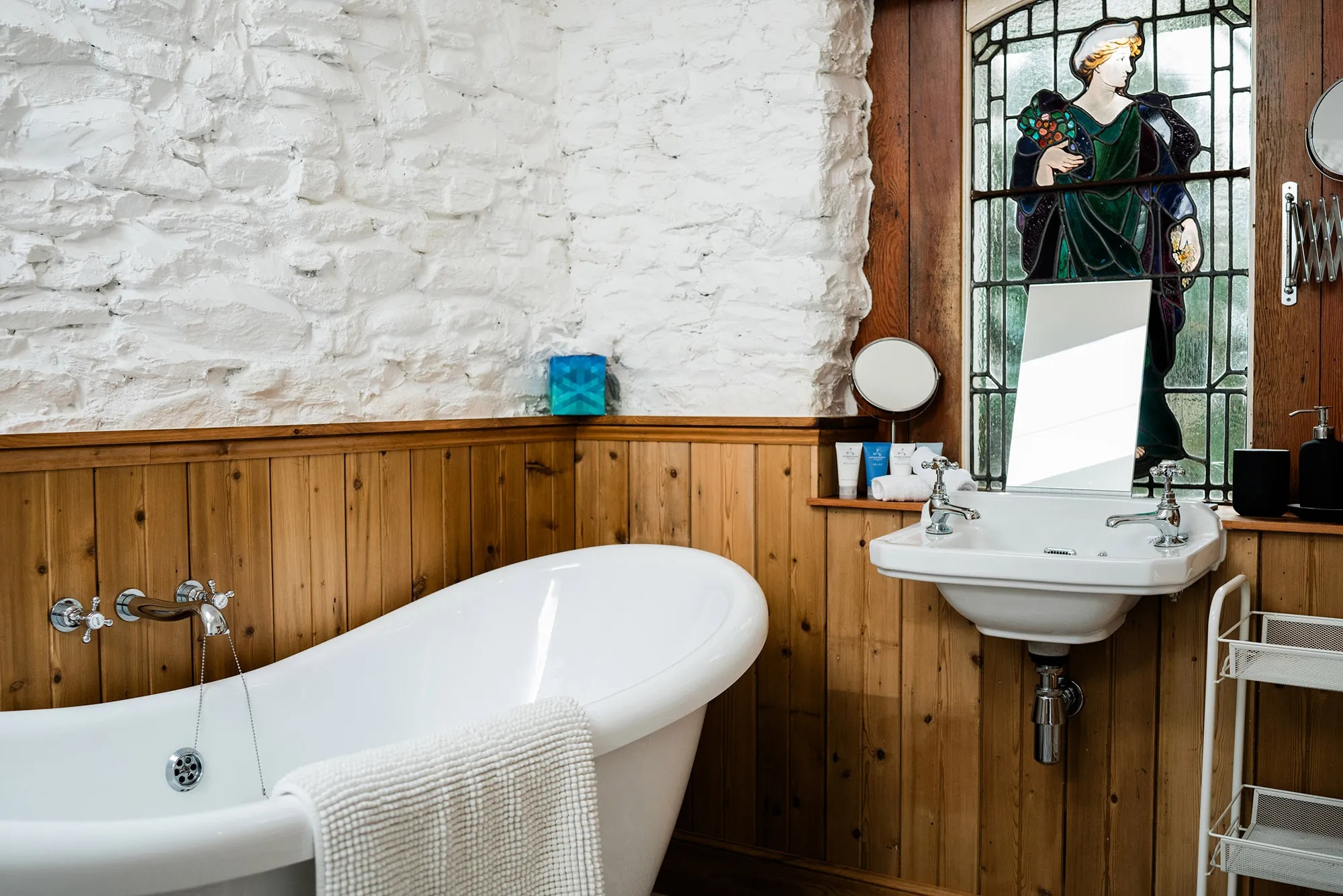 A roll-top bath stands against a whitewashed stone wall, a stained glass window and pedestal basin with traditional taps to the side.