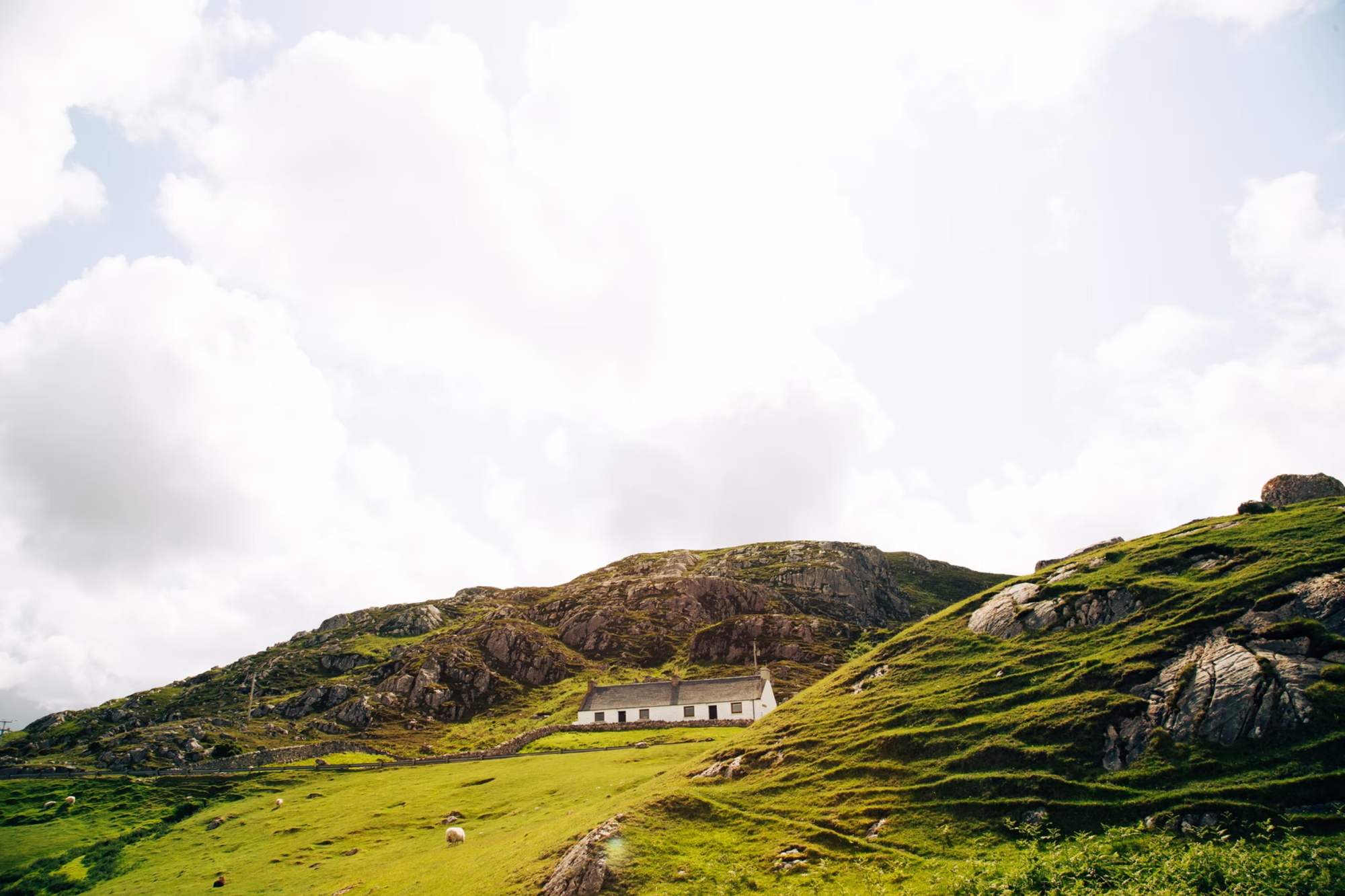A white church-like building sits at the foot of a rocky, grass-covered hillside, sheep grazing on the slope above.