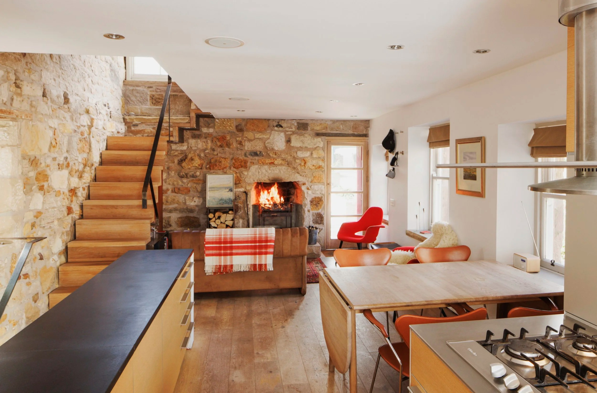 A wood burning stove is lit in a stone fireplace beneath a timber staircase, a dining table with orange chairs to the right and a red armchair beyond.