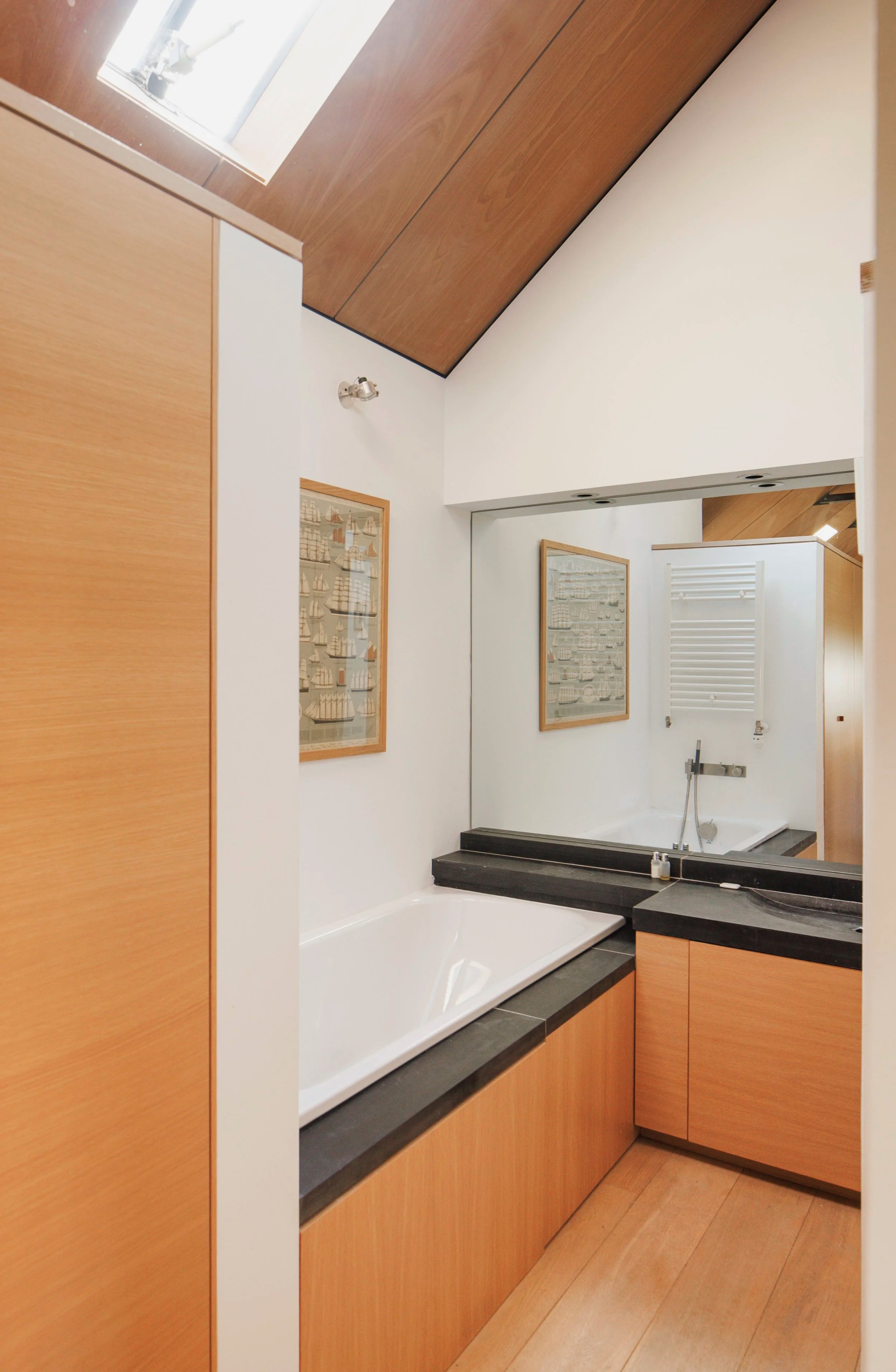 A bath with a black surround sits below a roof window, a wood-panelled wall and mirrored splashback reflecting the room.