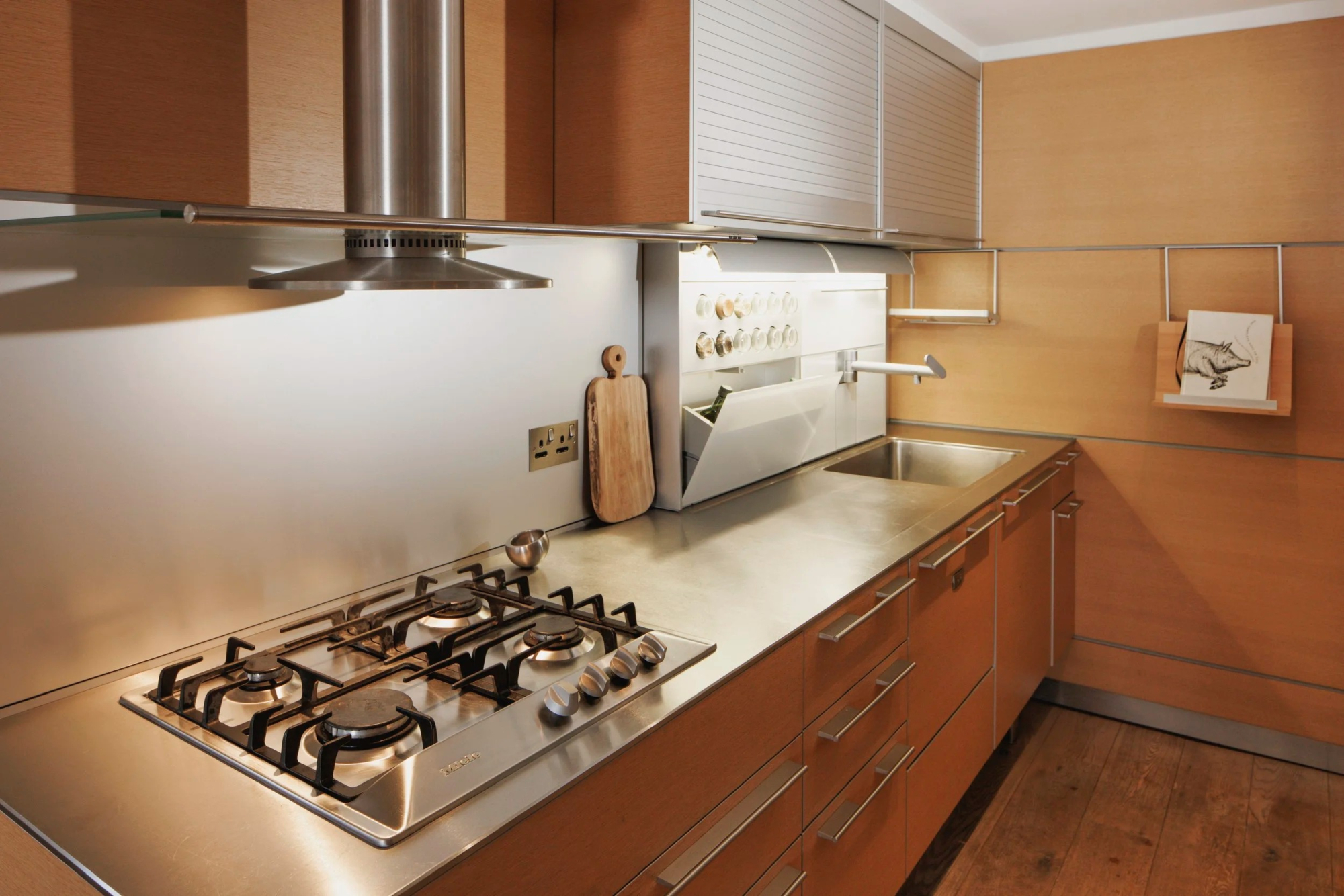 A gas hob and extractor hood stand in a warm wood kitchen, a chopping board and small ornaments on the stainless steel worktop.