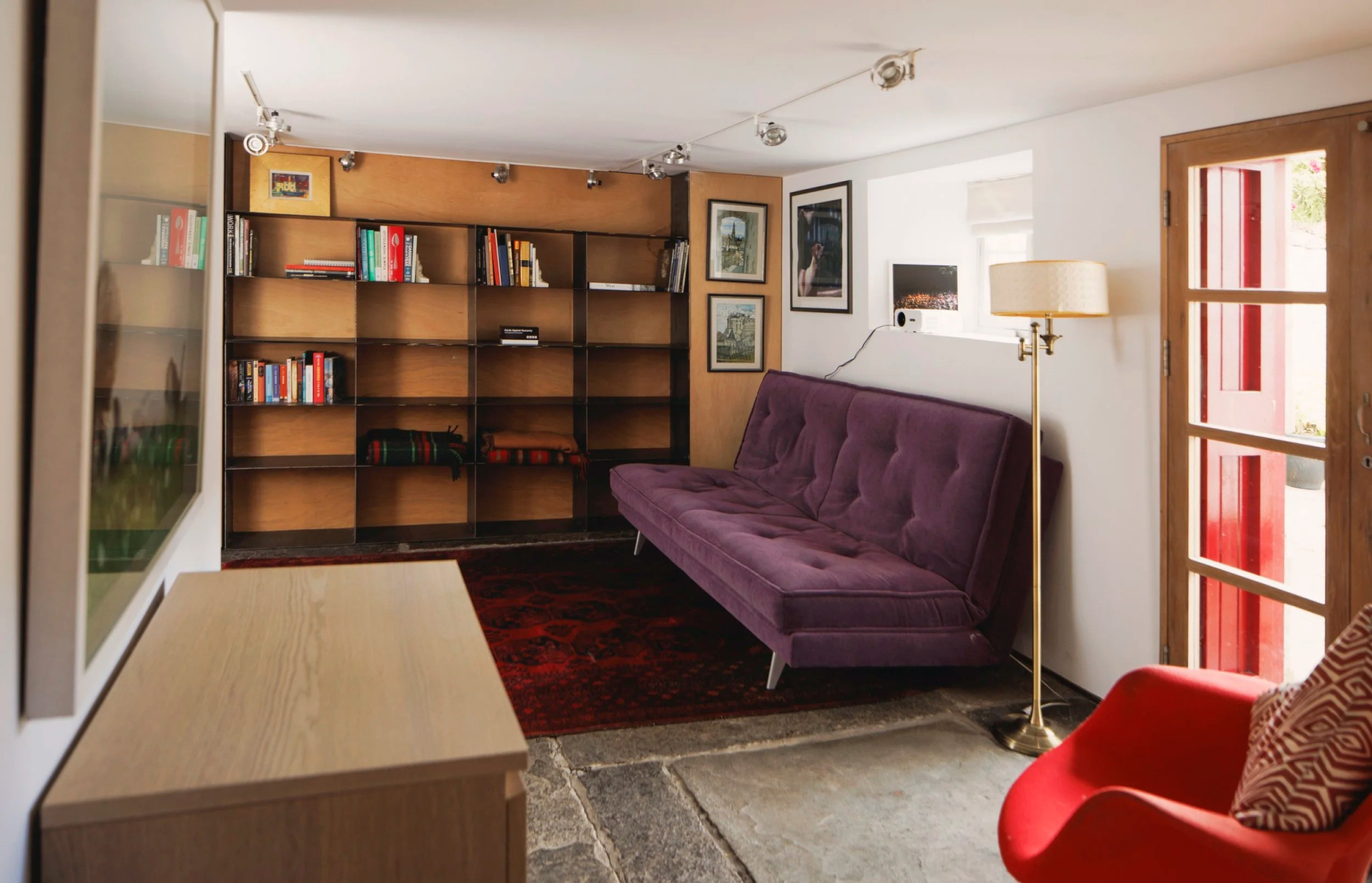 A purple velvet sofa and red armchair sit before built-in bookshelves, a red-framed glazed door opening to the side.