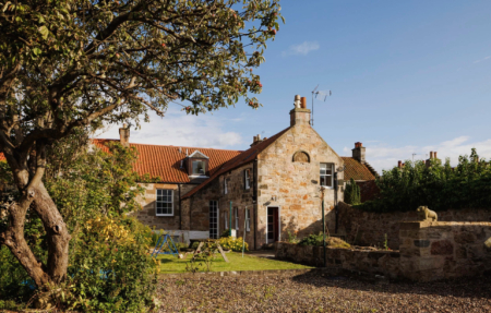 A stone cottage with red pantiled roofs and a gabled dormer sits behind a gravel courtyard, an apple tree in the foreground.