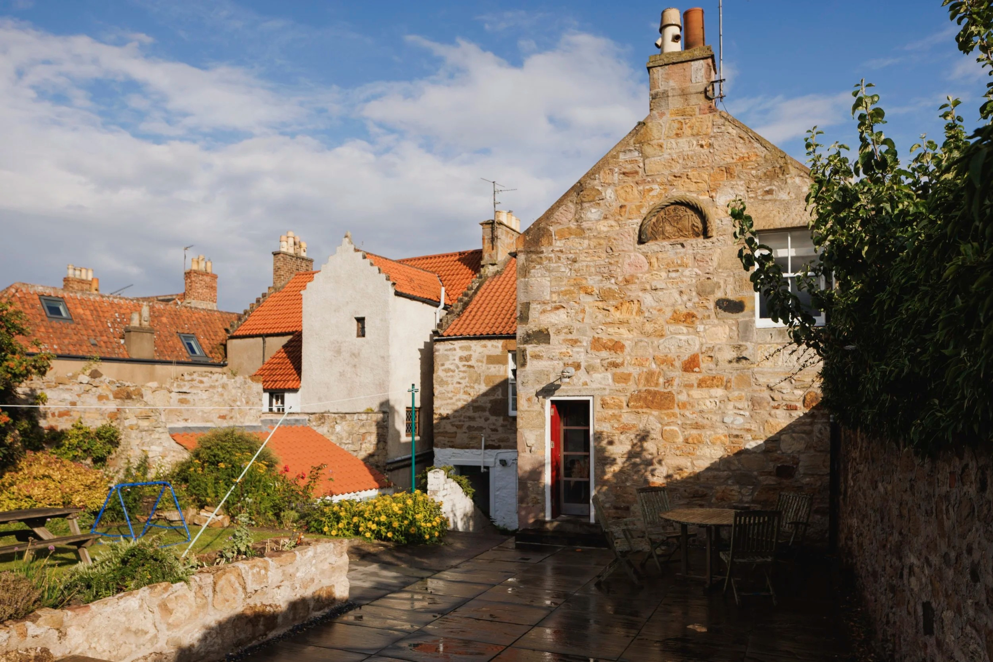 A stone cottage with a red pantiled roof and arched window detail sits behind a paved courtyard, a table and chairs set outside.