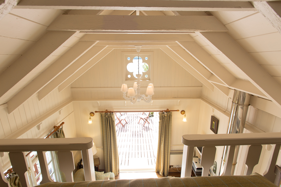 A vaulted timber ceiling with exposed beams rises above a mezzanine landing, a chandelier and curtained glazed door visible in the room below.