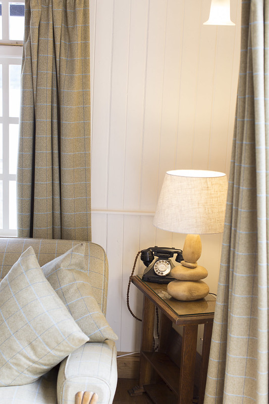 A white metal chandelier with lit lampshades hangs before a clover-shaped window looking onto a loch and hills.