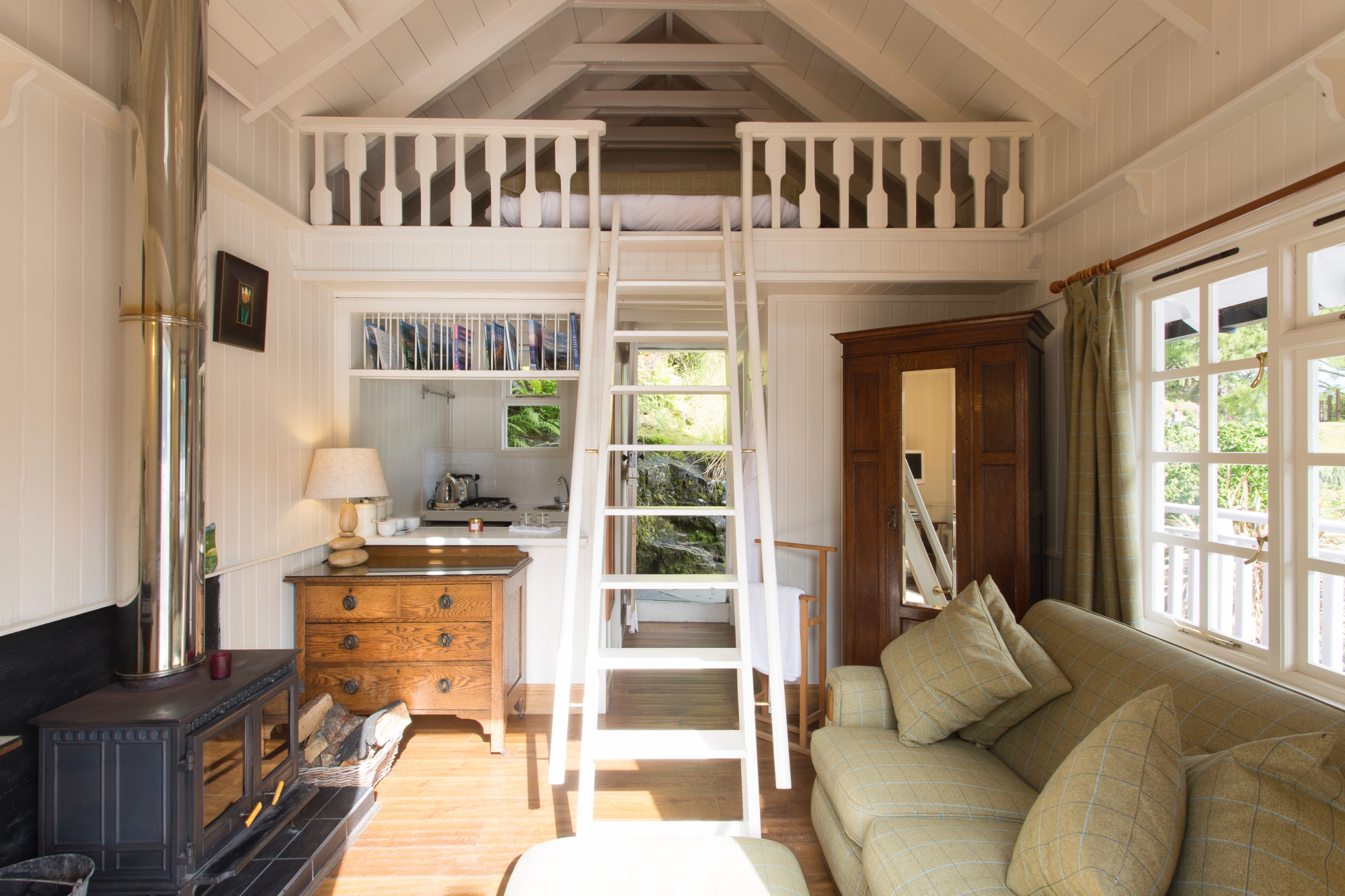 A wooden ladder leads up to a railed mezzanine loft, a wood burning stove, chest of drawers and cream sofa filling the room below.