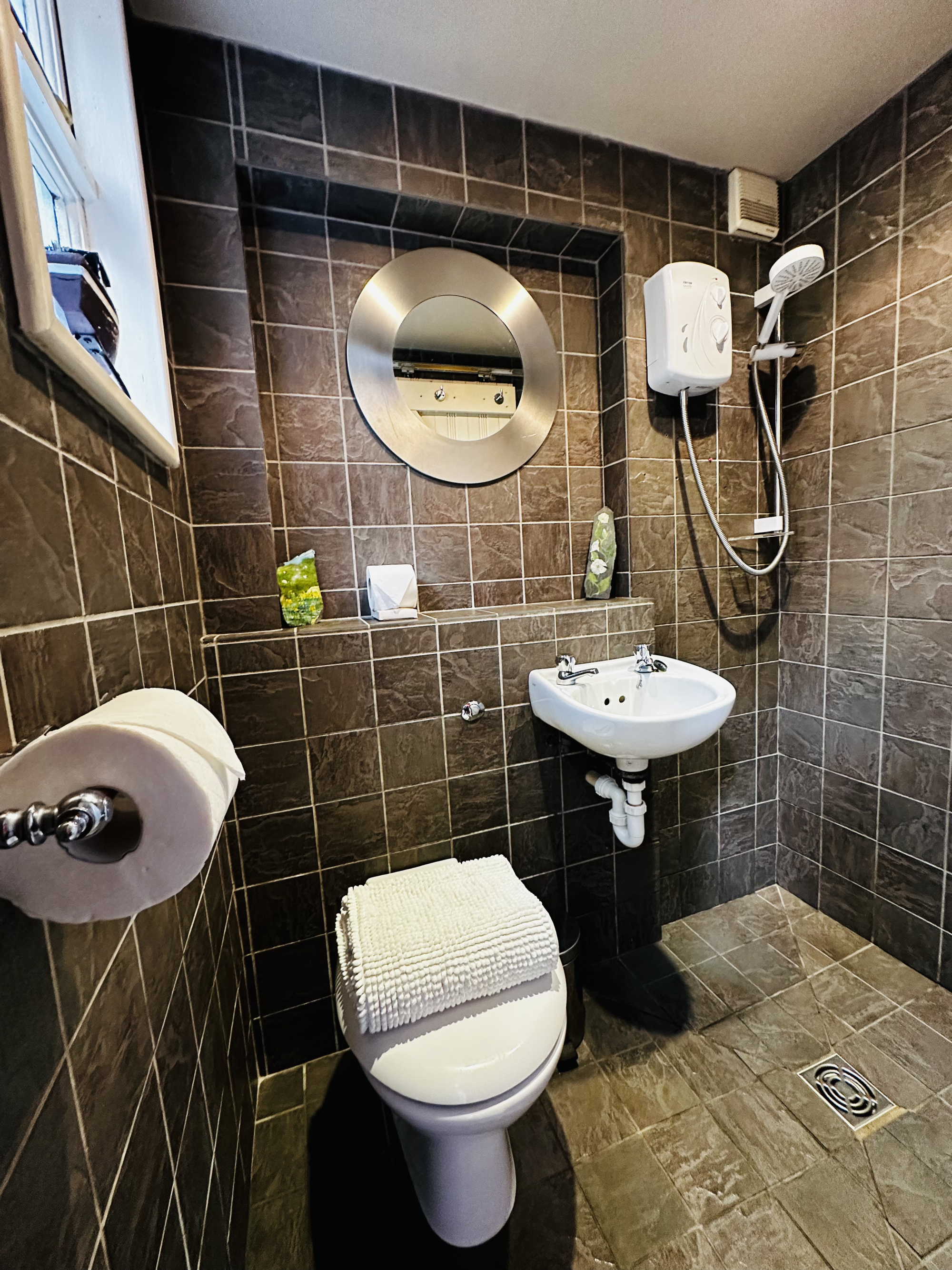A pedestal basin and toilet with a folded towel sit in a dark tiled bathroom, an electric shower and round mirror on the wall.