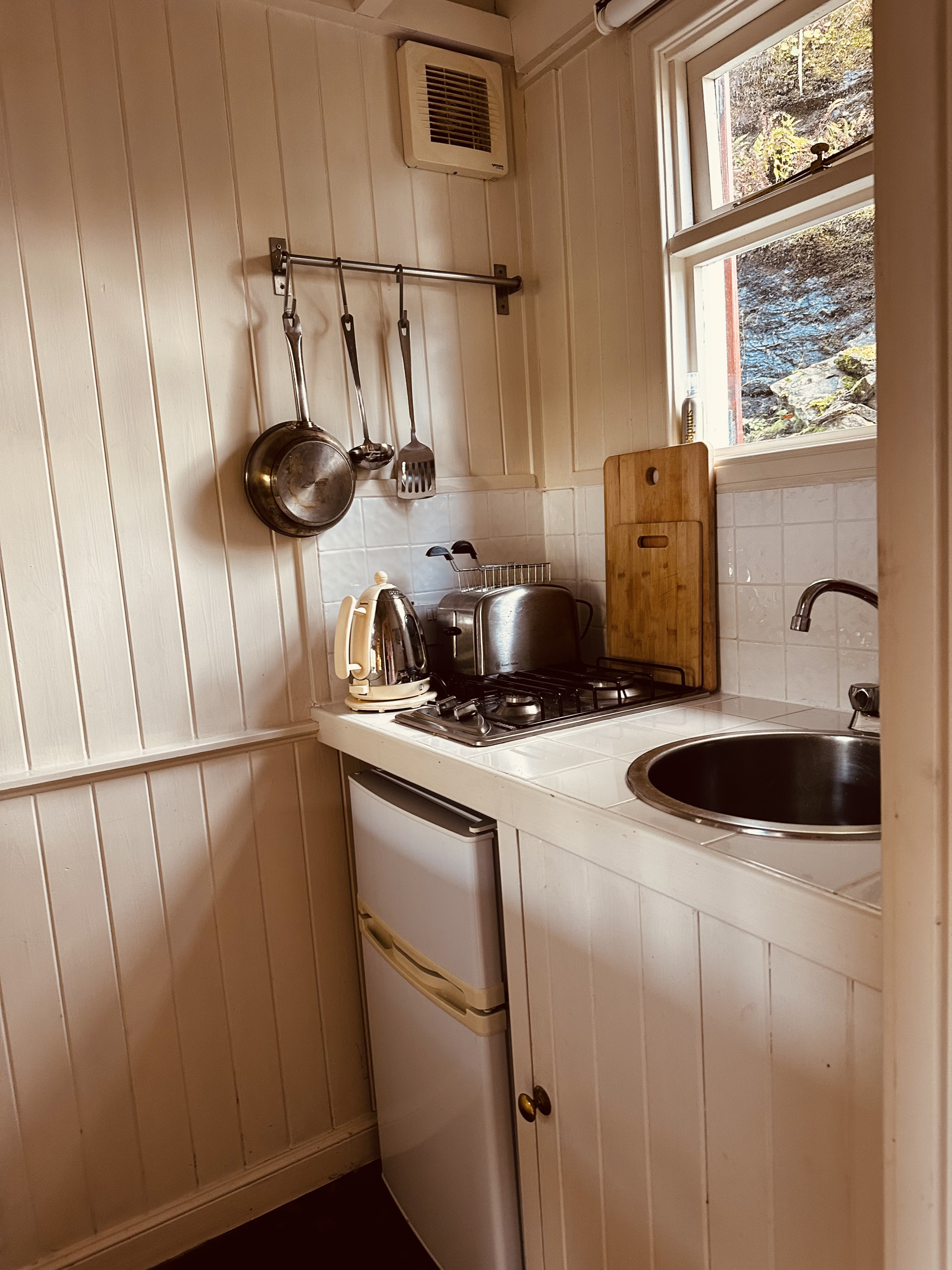 A small kitchenette with timber panelling holds a gas hob, round sink and hanging pans, a window looking onto rocks to the side.