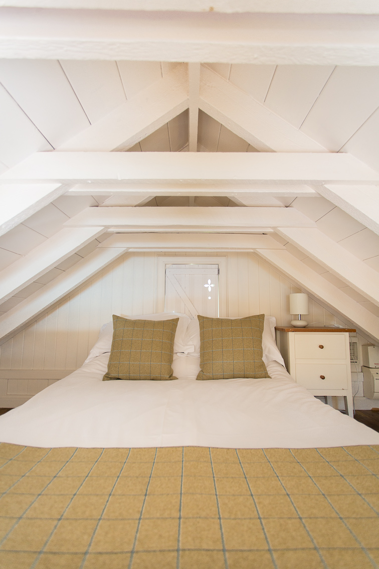 A vaulted timber ceiling looks down over a mezzanine balcony onto a room below, a chandelier and glazed door with striped curtains visible.