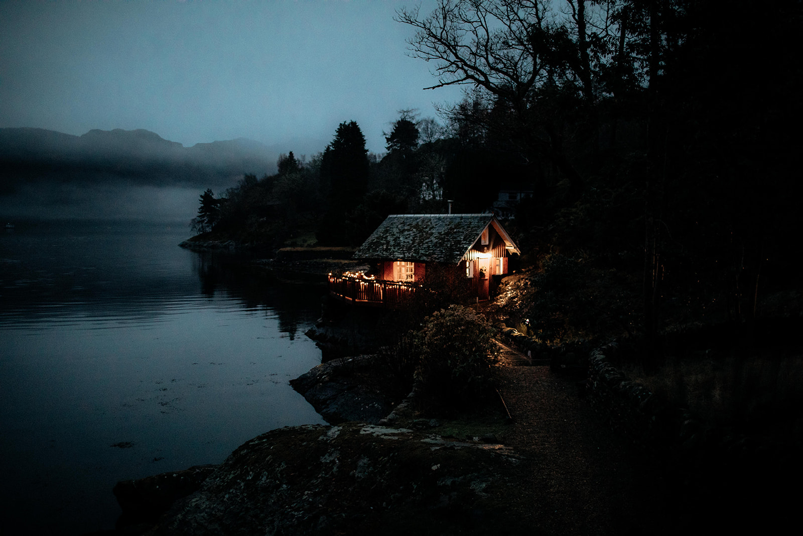 A timber lodge glows with lit windows on a loch shore at dusk, mist settling over the still water and hills.