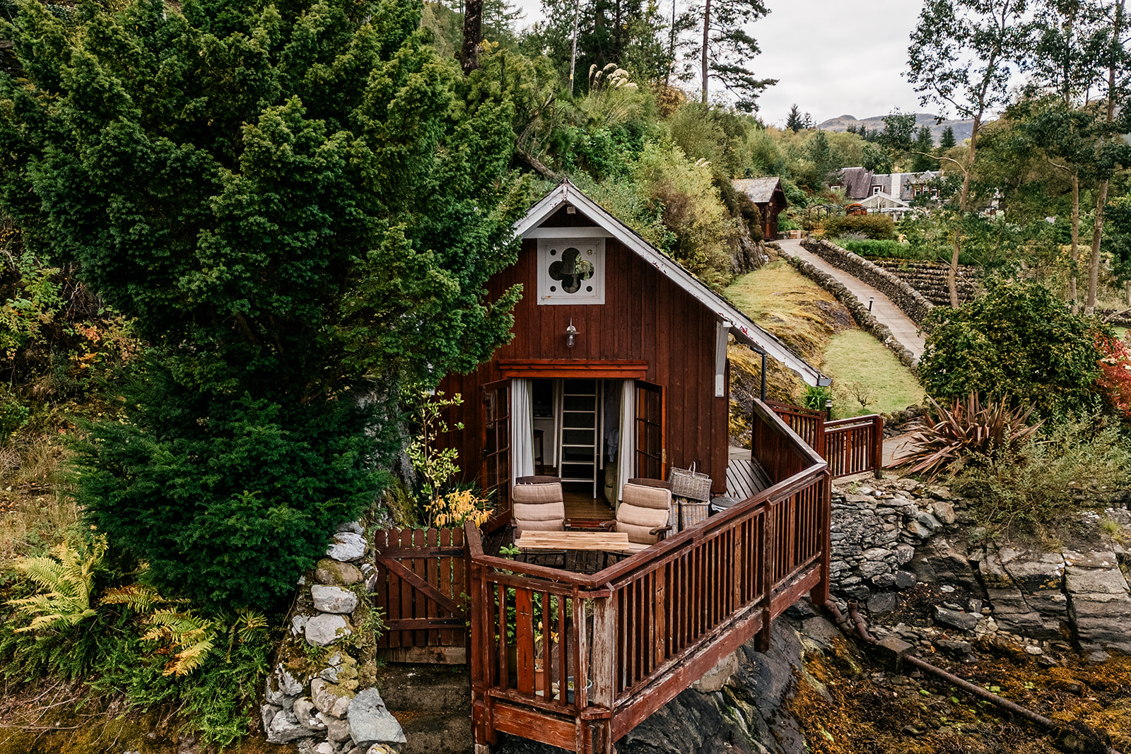 A dark timber cabin with a turf roof and carved gable end sits on a deck overlooking rocky woodland, chairs set outside the glazed door.